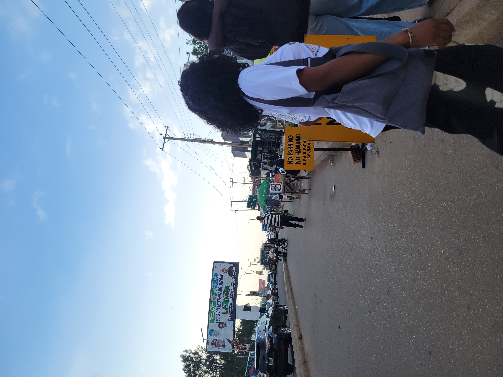 The image depicts a street scene in Jos, Nigeria, during what appears to be late afternoon or early evening, indicated by the warm lighting and the color of the sky. The foreground is dominated by a road with a slight curve, and a paved sidewalk on the right.
On the sidewalk, a young woman with dark, curly hair is walking away from the camera, wearing a white t-shirt and black pants with a facial print. She is carrying a gray shoulder bag. To her right, another person, partially visible, is wearing a dark top and blue jeans. Further to the right, a yellow sign with black text reads "NO PARKING NO HAWKING." Behind this sign, there's a stall or vendor setup with an umbrella and goods on display.
In the middle ground, several vehicles are present on the road, including a black SUV on the left, other cars, and a motorcycle with riders. A man in a black and white striped shirt is walking on the sidewalk towards the right. On the left side of the road, a large billboard displays political advertisements with images of individuals and text that reads "THE TIME IS NOW 2077 LET'S DO MUTFWANG AGAIN LE DMA."
The background reveals more of the urban environment with buildings, trees, and utility poles with overhead wires. The sky above is a clear blue with some scattered white clouds. The overall impression is that of a busy urban thoroughfare with commercial activity and political messaging.