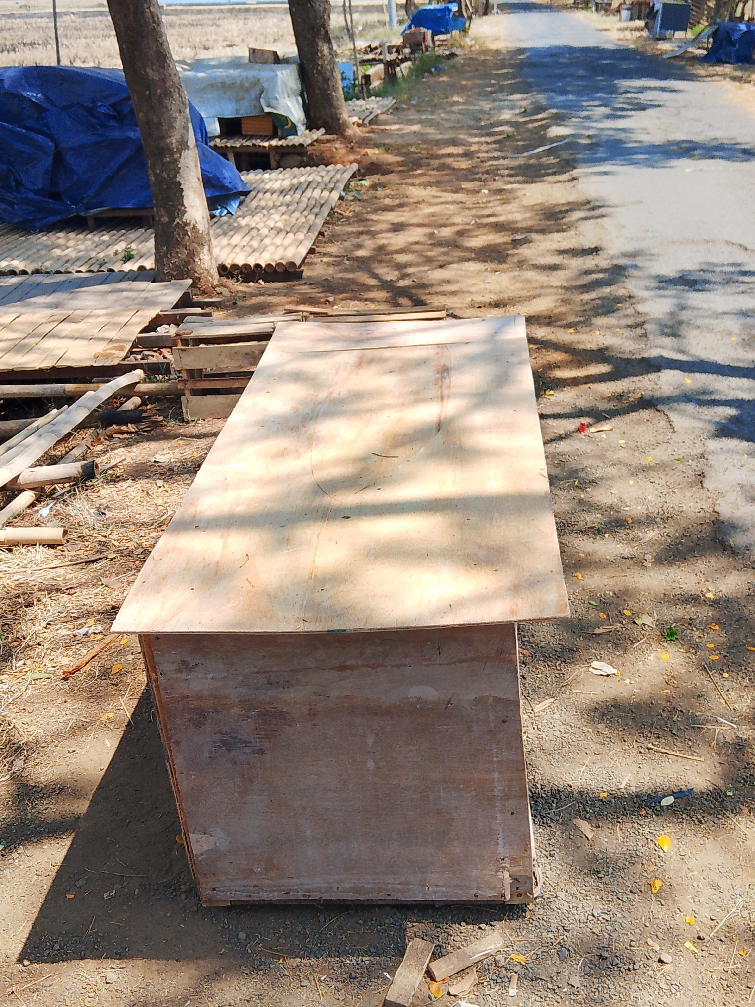 The image depicts an outdoor scene likely located in Kuanyar, Indonesia. In the foreground, a wooden box-like structure topped with a plywood sheet is positioned on the ground, which is a mix of dirt and gravel. Sunlight casts dappled shadows from trees onto the structure and the ground. To the left of the structure, several wooden planks and bamboo poles are visible, along with a large blue tarp spread over some other objects. In the background, a paved road stretches into the distance, lined with trees and vegetation. The overall impression is of an informal outdoor storage or workspace, possibly related to construction or local trade.