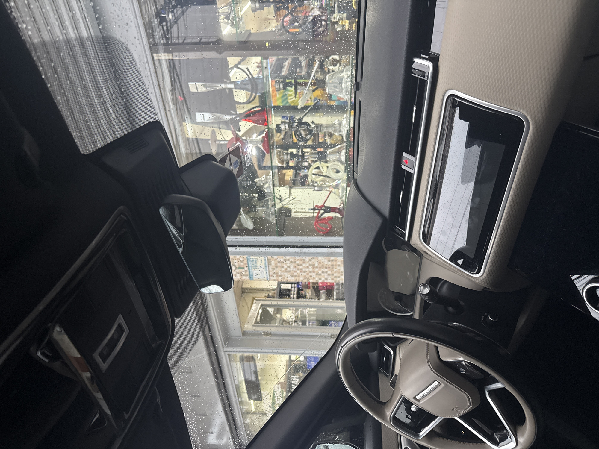 The image shows the interior of a car, specifically the dashboard, steering wheel, and windshield. The car is parked in front of a building that appears to be a workshop or store, as indicated by the display of various mechanical parts and tools visible through the rain-streaked windshield. The general view outside does not allow a definitive location in Mexico City to be ascertained.