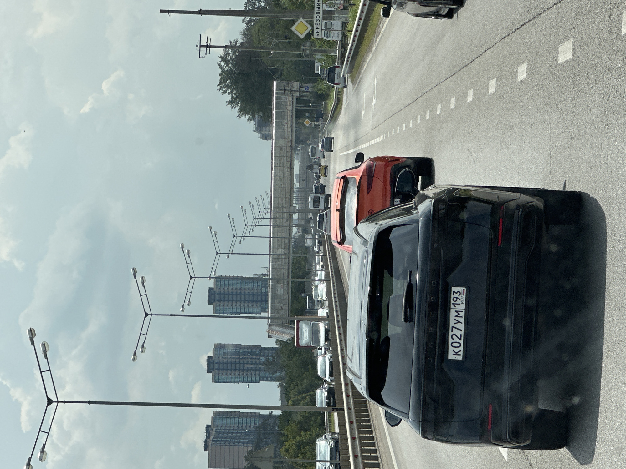 The image shows a traffic jam on a highway, likely within the city limits of Beryozovy, Russia, given the signage. In the foreground, a black SUV is positioned in the left lane, its rear visible to the viewer. The license plate reads "K027YM 193 RUS". To the right and slightly ahead of the black SUV is a bright orange sports car. Several other vehicles, including vans and cars, are visible further down the highway, indicating moderate to heavy traffic.
Overhead, multiple streetlights with multiple bulbs are mounted on tall poles. In the middle ground, a pedestrian bridge crosses the highway, with a bus and other vehicles visible beneath it. Beyond the highway, residential buildings with a distinctive architectural style, likely apartment complexes, can be seen in the distance. Green trees and foliage line the sides of the road and surrounding areas. The sky is partly cloudy, suggesting overcast weather. A road sign with a yellow diamond shape and Russian text "БЕРЁЗОВЫЙ" (Beryozovy) is visible on the right side of the road, confirming the location. The overall scene depicts a typical urban highway environment during daylight hours.
