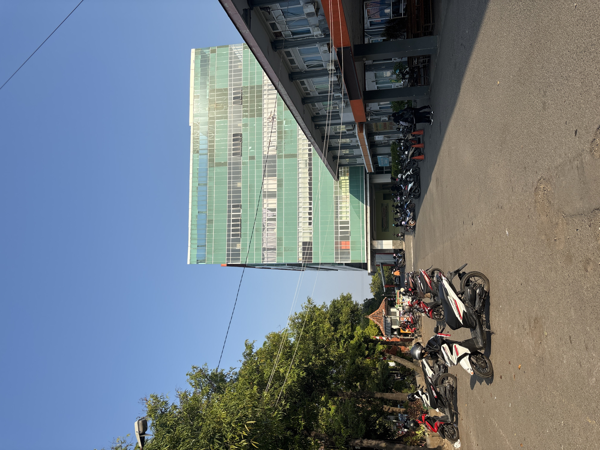 The image depicts the exterior of a modern, multi-story building with a green glass facade, likely a hospital or office building, situated in a city environment. The building appears to be a prominent structure in the scene. In the foreground, several motorcycles are parked, suggesting a public or commercial area. A row of trees lines the left side of the image, providing a natural element to the urban landscape. The sky is clear and blue, indicating a sunny day. The architecture suggests a location in Indonesia, and further research confirms this building as the RSU Gunung Jati Hospital in Cirebon, Indonesia, a well-known public hospital in the city. The image captures a typical day with people coming and going, indicated by the parked vehicles and the presence of individuals walking in the distance. The overall atmosphere is one of an active, functional urban space.
