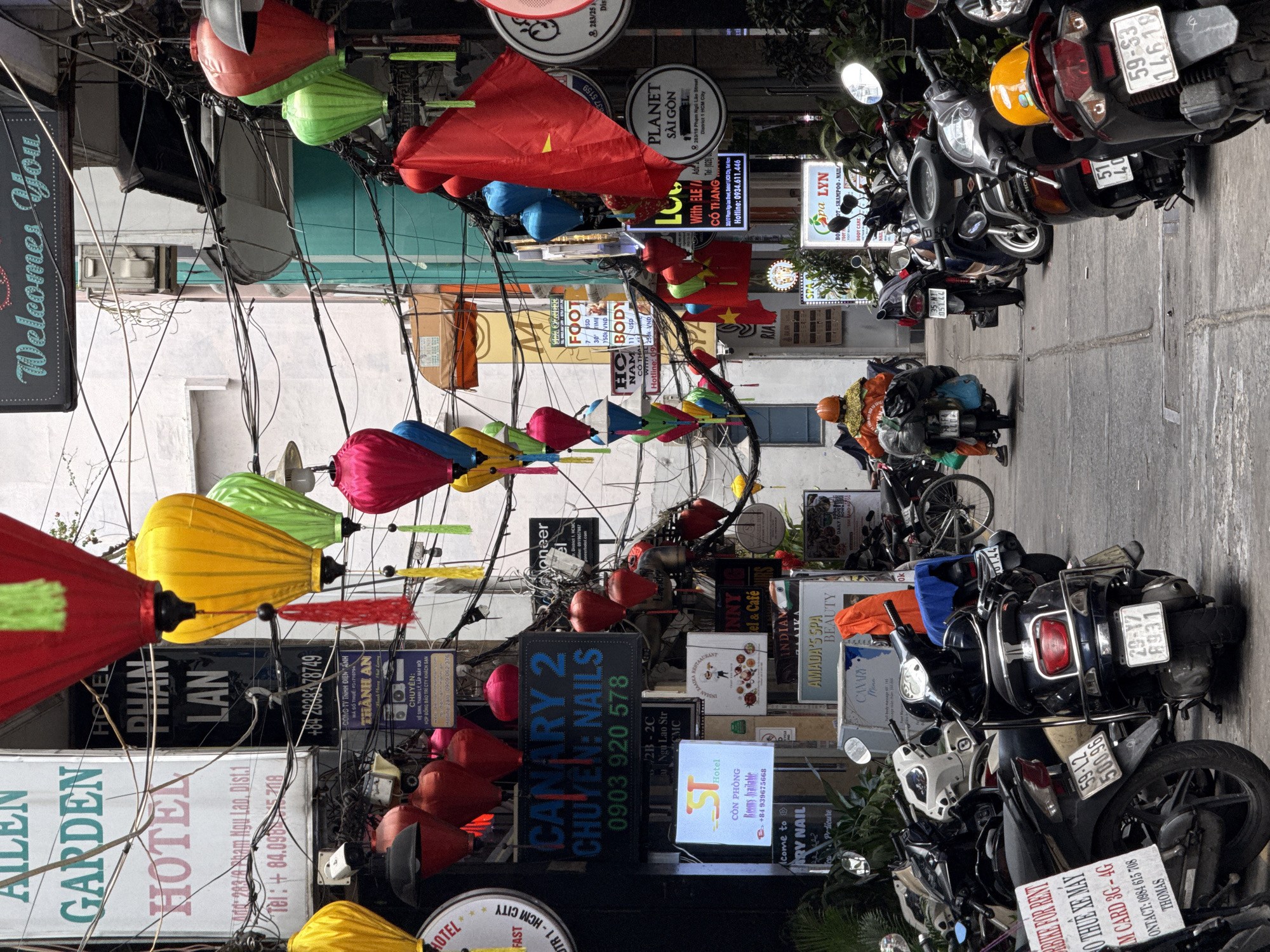 The image shows a street scene in a Vietnamese city, likely not Thủ Đức given the presence of Phạm Ngũ Lão Street, which is located in District 1, Ho Chi Minh City. The street is crowded with parked motorbikes on the right side. Overhead, there is a dense network of electrical wires and colorful lanterns strung across the street. Numerous signs advertising businesses, including hotels, spas, restaurants, and nail salons, are displayed. A person wearing an orange helmet and heavily laden with bags is seen walking on the street. The overall impression is of a busy, vibrant urban environment.
