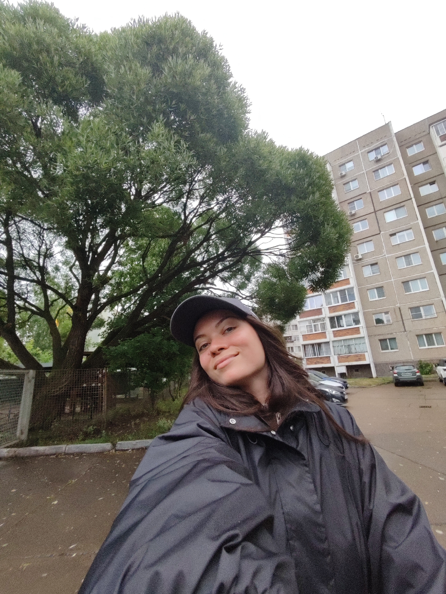 In an outdoor, urban setting, a young woman with long, dark brown hair and a grey baseball cap is taking a selfie. She is wearing a dark grey, possibly black, hooded jacket. Her head is tilted slightly to her right, and she is smiling. Behind her, on the left, is a large tree with dense, green foliage. To the right of the tree and extending into the background is a tall, beige apartment building with multiple floors and numerous windows. Several cars are parked in a lot in front of the building. The ground is paved and appears wet, with scattered leaves. The sky is overcast. This scene is consistent with a residential area in the city of Chaikovsky, Russia, likely in a courtyard or parking lot adjacent to an apartment complex.