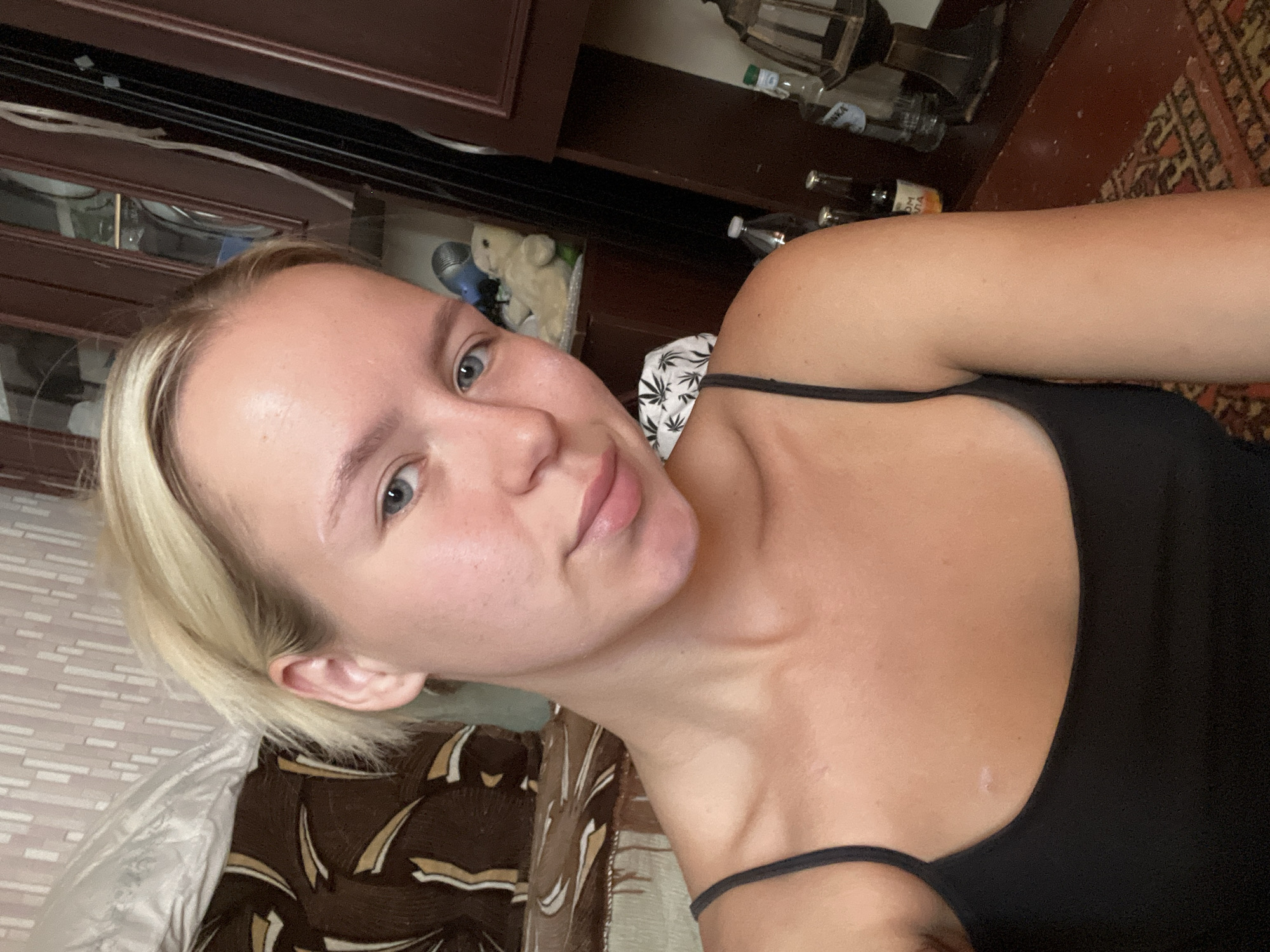 A young, fair-skinned woman with blonde hair tied back in a messy bun is taking a selfie. She is wearing a black tank top. Her eyes are blue and she has a neutral expression. The background appears to be a room with patterned wallpaper and wooden furniture. On a shelf behind her, there is a small stuffed bear and a blue object that might be a speaker. On a surface in the foreground, there are several glass bottles and a lamp. The scene is dimly lit. The location is described as Unknown, Unknown.