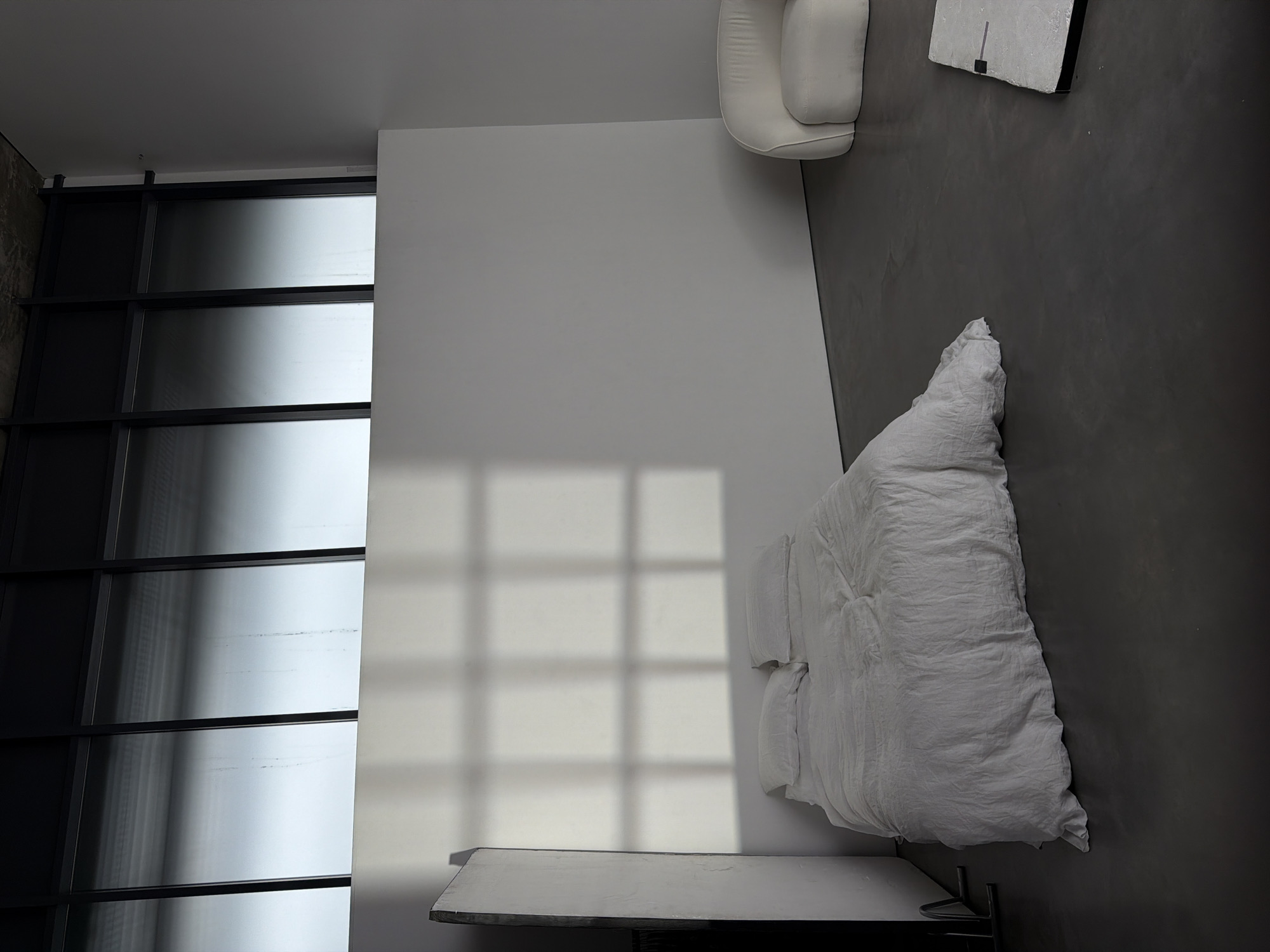 An overhead, interior shot of a modern minimalist bedroom, possibly in London, UK. A large window with black framing takes up the left side, casting grid-like shadows on the white wall beside it. A white bed with a rumpled duvet and pillows is situated on the right, partially on a dark grey floor. A small white armchair is in the upper right, and a modern, rectangular object is adjacent to it. A white bedside table sits next to the bed on the lower left side. The room appears stark and devoid of people or activity.