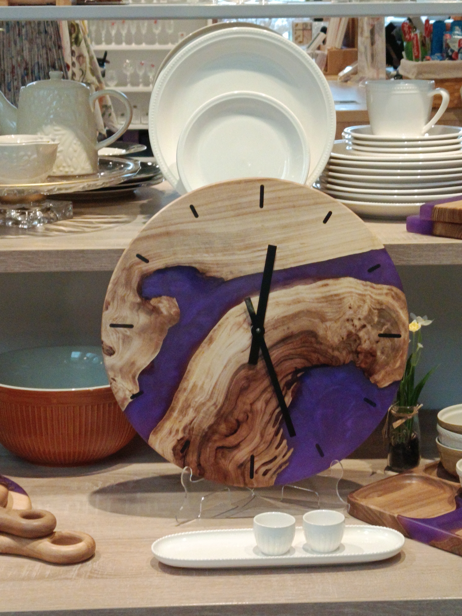 The image depicts a still life arrangement of various household items on a wooden shelf or table. The items include white dishes (plates and a teapot), a round wooden clock with a purple resin inlay, a brown ceramic mixing bowl, wooden serving utensils, a small vase with flowers, and two white ceramic cups on a small white serving dish. The objects are arranged in a way that suggests they are on display, possibly in a retail setting or a home decor arrangement. The background includes additional glassware and dishes on shelves. Based on the style of the items and the context of the image, it is plausible that this scene could be located in a home goods store or artisan market in Moscow, Russia, or any other city with similar consumer goods and decorative items.