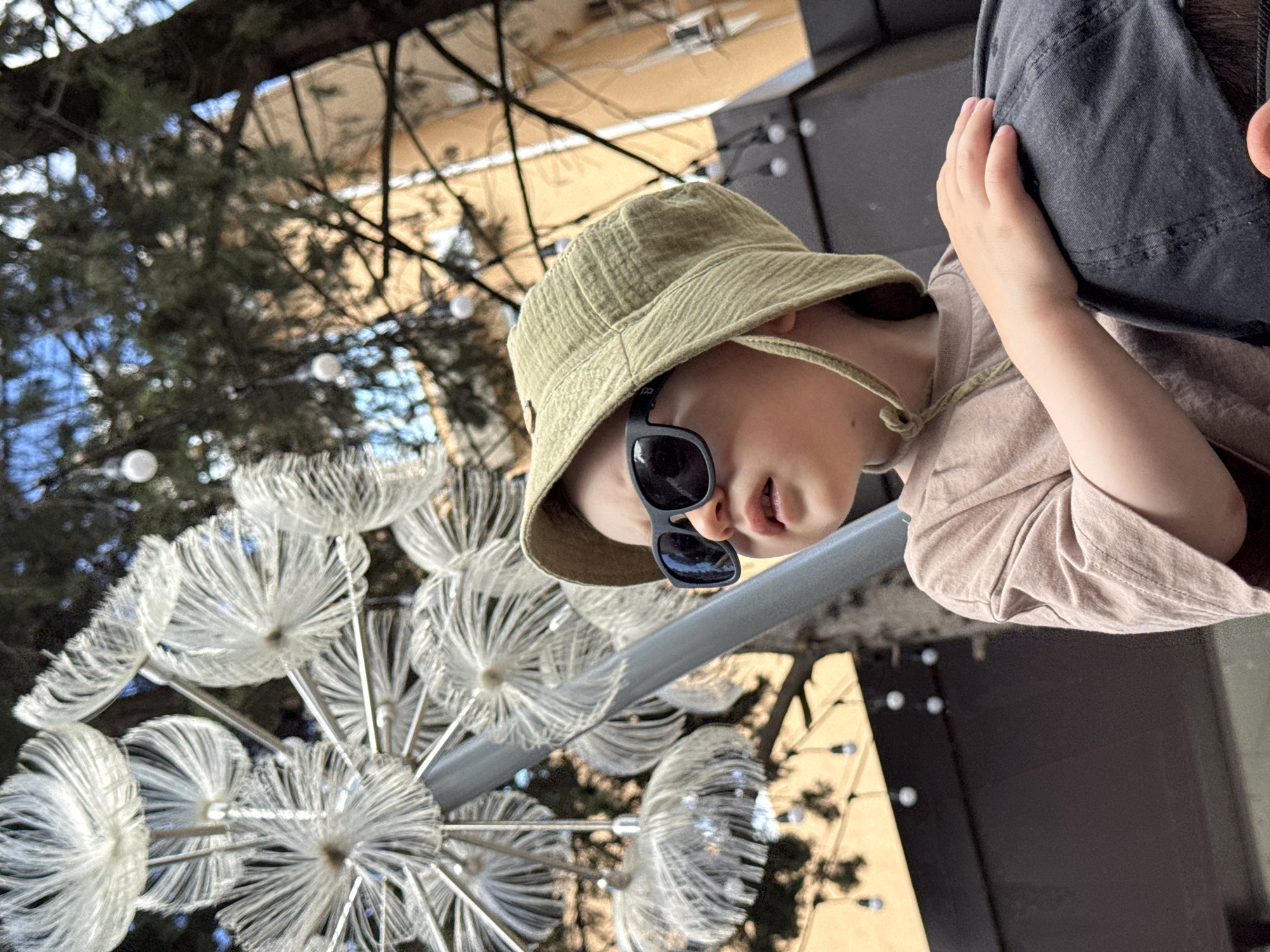 A child wearing a tan hat and dark sunglasses is positioned near an elaborate, multi-tiered dandelion-like light fixture. The child is wearing a tan shirt and is close to a person wearing a dark garment. The scene appears to be outdoors, under some trees. The light fixture and trees hint at a public space within a city.
