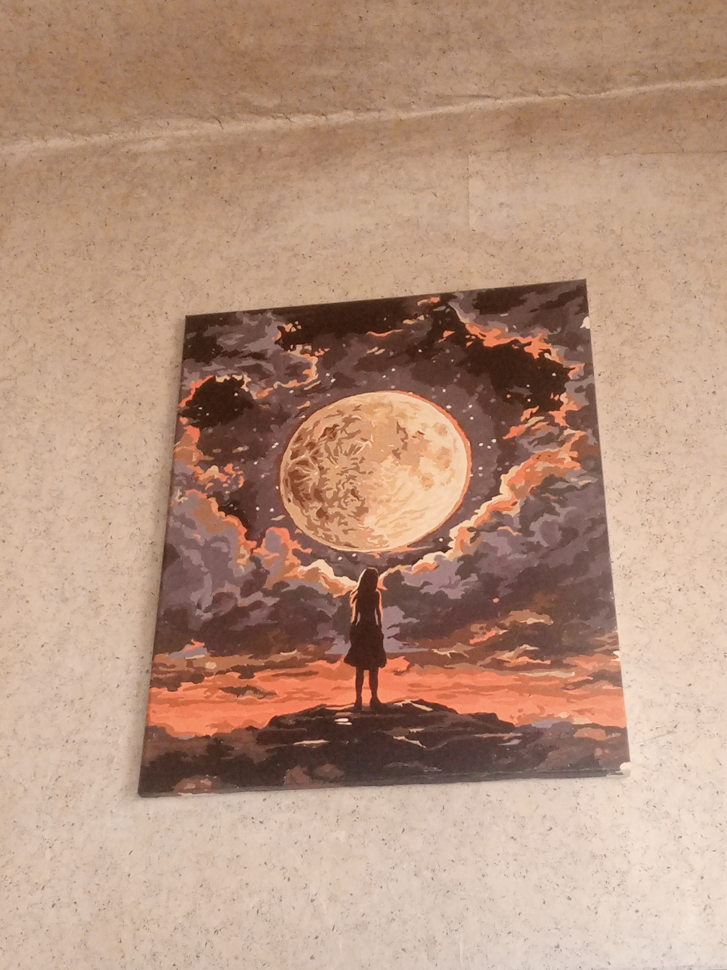 The image depicts a painting of a person standing on a rocky outcrop, gazing at a large, textured moon in a cloudy, star-filled sky. The sky is a mix of dark purples, grays, and fiery oranges, suggesting either a sunset or an otherworldly celestial event. The moon dominates the upper portion of the canvas, its surface rendered with detailed shading. The figure is silhouetted, wearing a long dress, and appears small in comparison to the vastness of the sky and moon. The painting is displayed on a wall with a textured, light-colored surface, possibly a stucco or concrete finish. The location is not identifiable from the image itself.