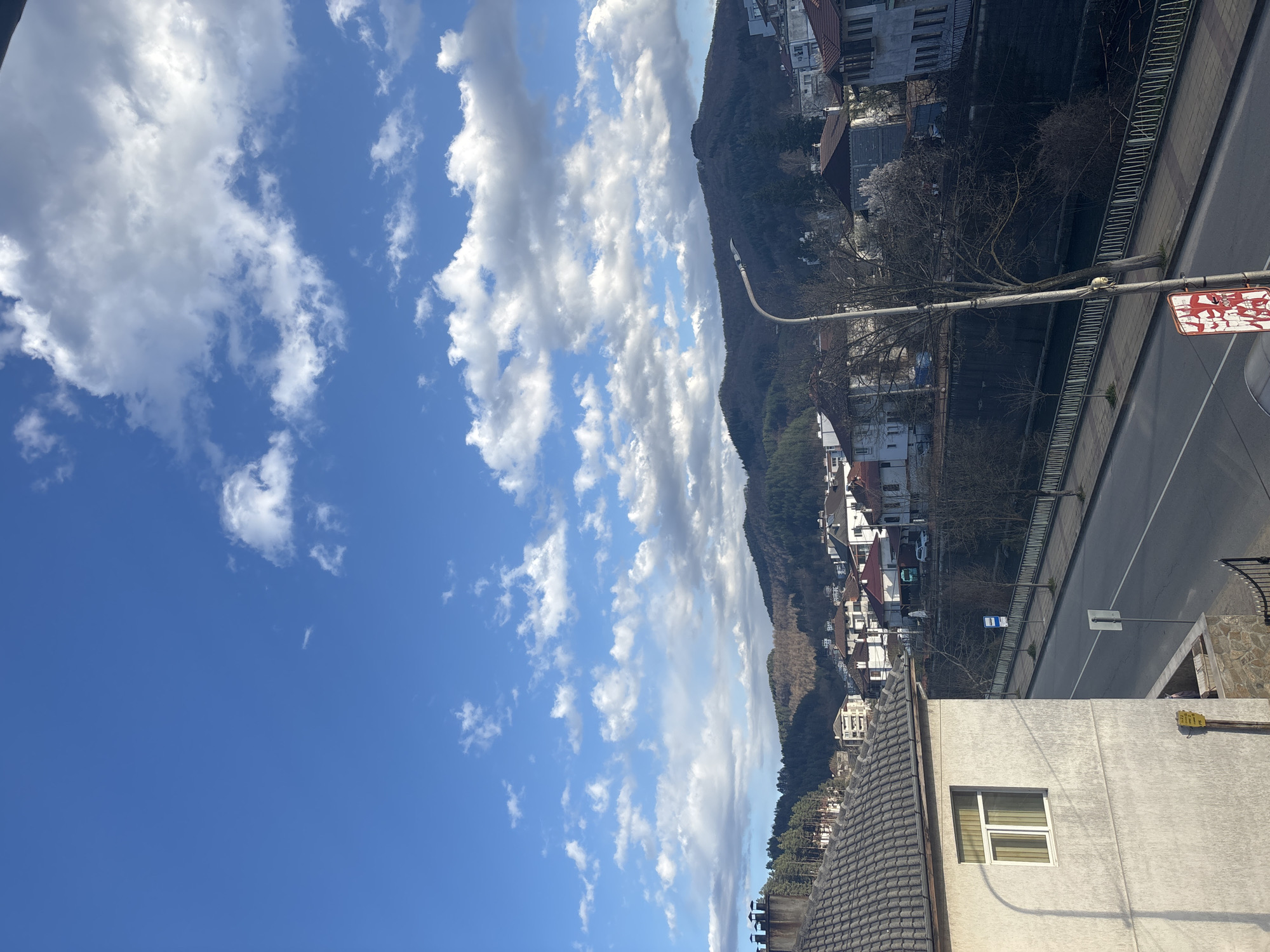 The image shows an elevated view from Zlatograd, Bulgaria. A clear blue sky with scattered white clouds is visible. In the background, a forested mountain rises. Closer to the camera, a street runs alongside a retaining wall. Buildings and houses are visible on both sides of the street, with some partially obscured by trees. In the foreground, a white building with a window and a gray tiled roof is visible. A streetlamp and a traffic sign stand beside the road.