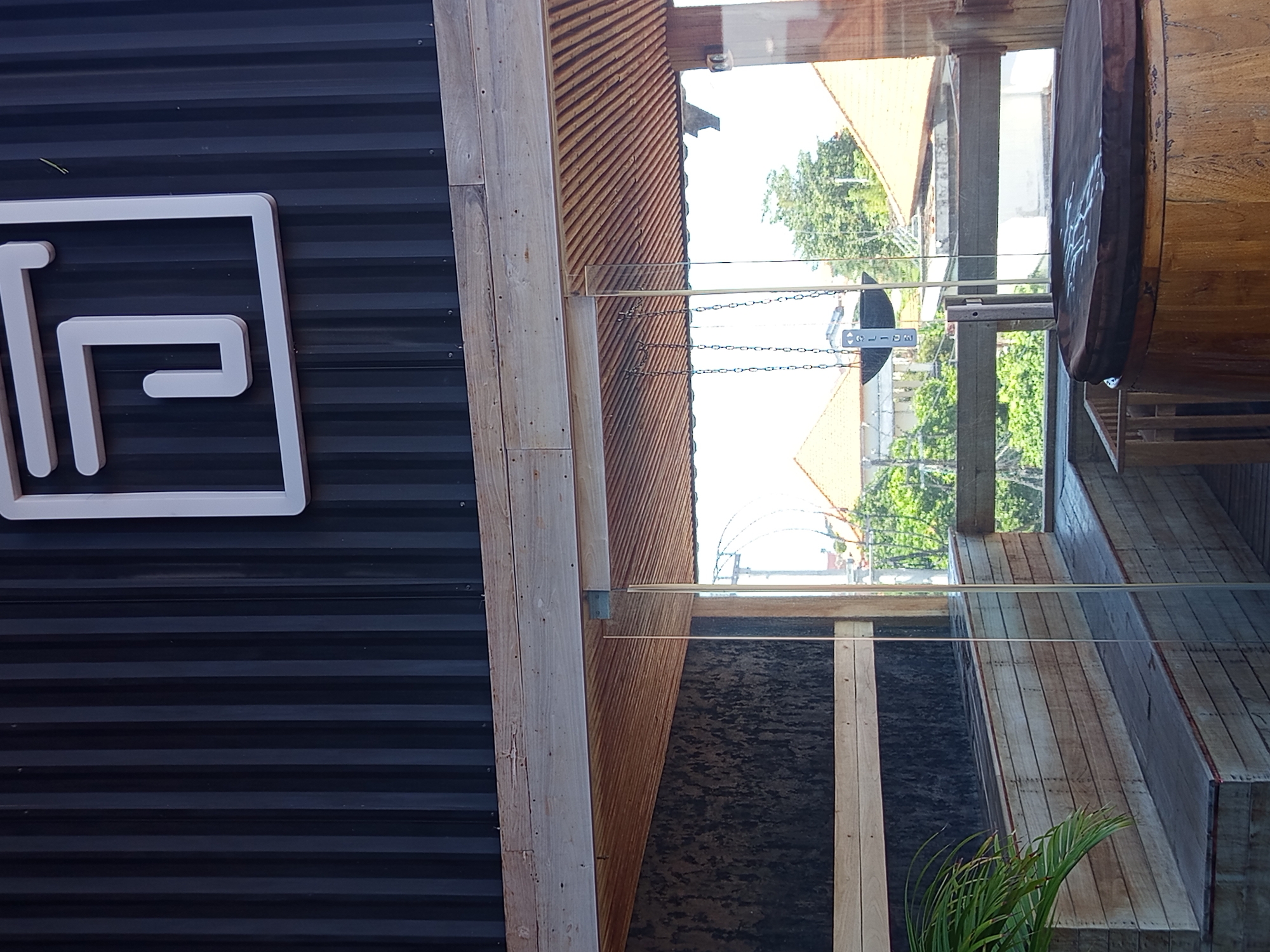 The image shows a partial view of a building exterior, possibly in North Kuta, Indonesia, judging by the tropical vegetation visible through a glass window. The left side features a dark, corrugated metal wall with a white, square logo that includes the letter "P." Adjacent to the metal wall is a wooden structure with a horizontal plank design. The wooden structure extends towards a glass window that reflects the exterior environment, including trees and buildings with orange roofs. Inside the building, as seen through the glass, there's a wooden barrel-shaped object and a multi-level wooden structure. The interior also contains a plant with green fronds. A hanging sign is suspended by a chain and is visible through the glass.