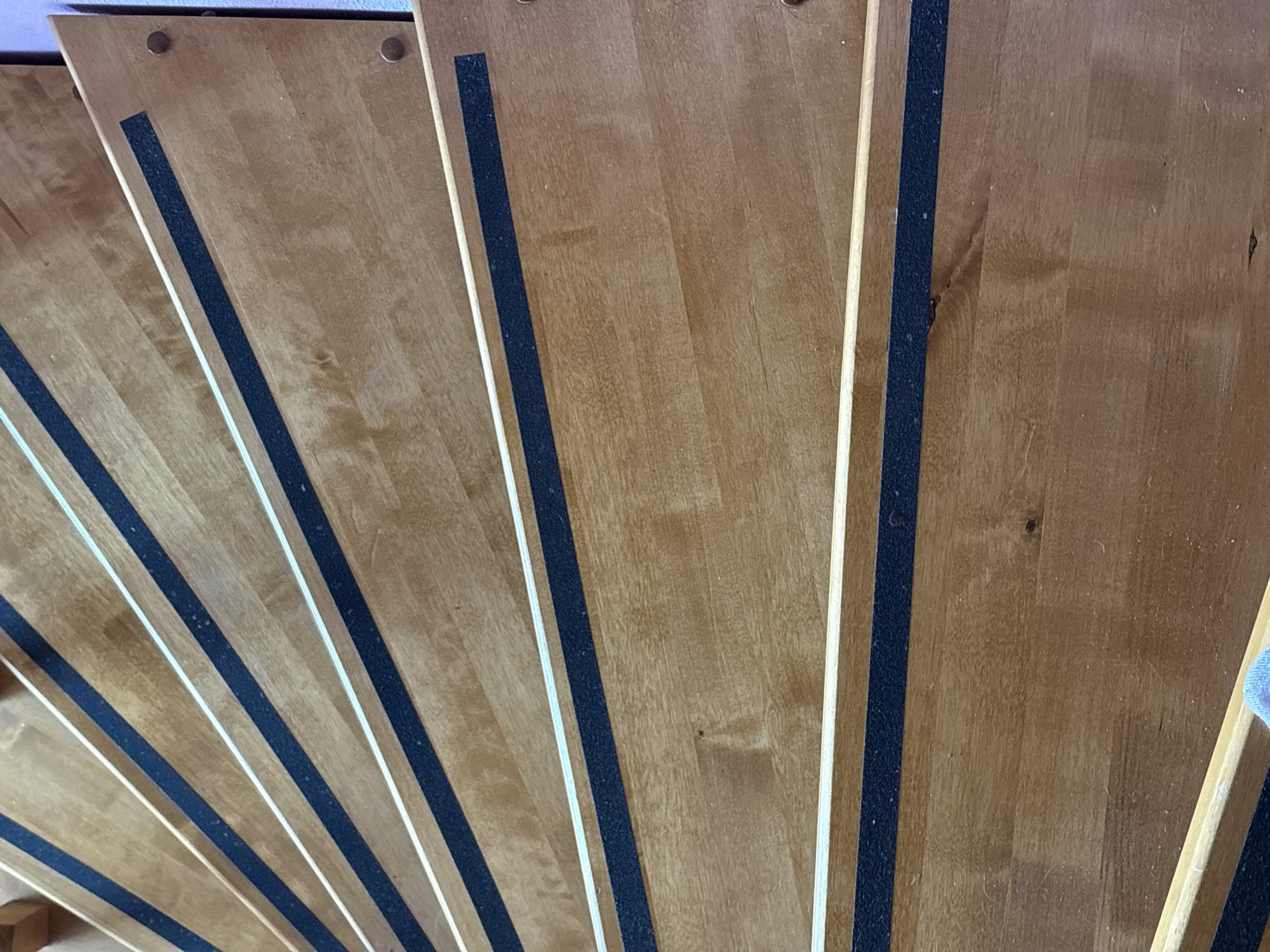 The image shows a close-up of winding wooden stairs. Black anti-slip strips are adhered to each stair tread. The wood appears to be light brown in color. The view is from above, looking down the staircase. There is no indication of people in the image, nor any other objects beyond the stairs and anti-slip strips. The location is presumed to be Прикамский, Russia, based on the user's request, but there is no discernible visual evidence in the image to confirm this.