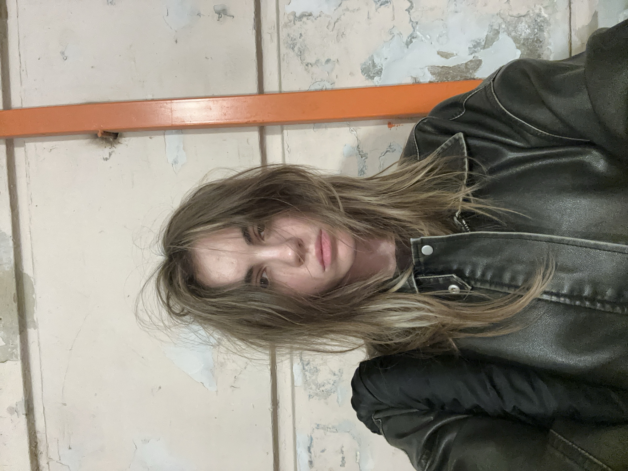 A young woman with messy brown hair and a black leather jacket is standing in front of a textured, peeling wall. An orange pipe runs vertically along the wall behind her. The lighting is dim and appears to be artificial. The woman is looking directly at the camera with a neutral expression.