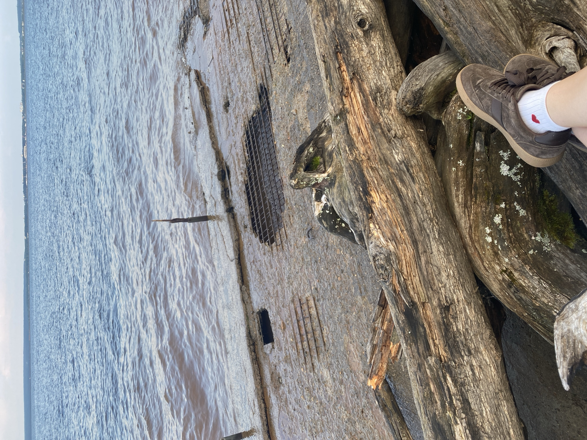 The image shows a person's legs and feet resting on a pile of driftwood logs near the shore. The person is wearing brown sneakers and white socks with a small red heart design on one sock. The logs are weathered and appear to be old. Behind the logs, there is a concrete structure partially submerged in the water, with metal grates visible. The water is choppy and a murky brown color, with the horizon visible in the distance under an overcast sky. The location appears to be the Kama River embankment in Chaikovsky, Russia, a city known for its industrial and hydroelectric power facilities, which could explain the presence of the concrete structure.