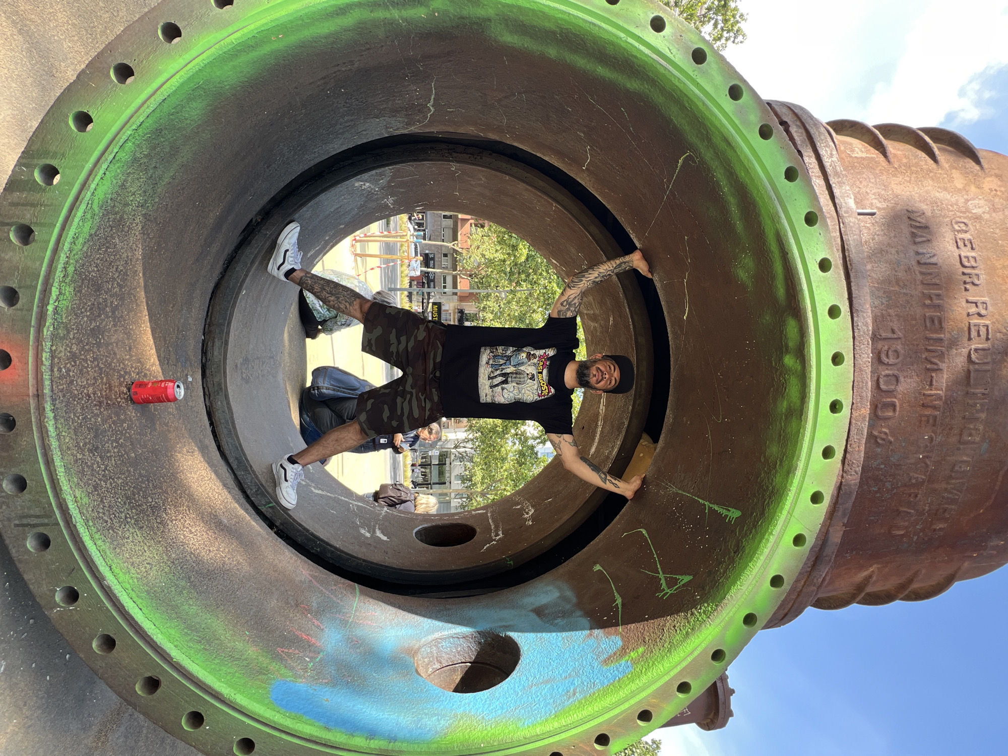 The image depicts a man positioned inside a large, horizontal pipe structure. The man is wearing a black t-shirt, camouflage shorts, white sneakers, and a baseball cap. He has tattoos on both arms and is smiling at the camera. He is holding on to the inner sides of the pipe with his hands and positioning his feet against the opposite side of the pipe.
The pipe appears to be made of metal, with a weathered exterior. It is painted with green edging and also has blue and green graffiti markings. The pipe has circular openings along the outer edge. A red beverage can sits on the pipe interior to the left of the man, next to a denim backpack.
Visible through the open ends of the pipe is an urban scene, including trees, buildings, street signs, and blurred human figures walking past. Based on visual cues and the description, it can be confirmed that this is the "L'Estació del Nord" sculpture park, which is located in Barcelona, Spain. This sculpture park features various large pipes as an art installation.