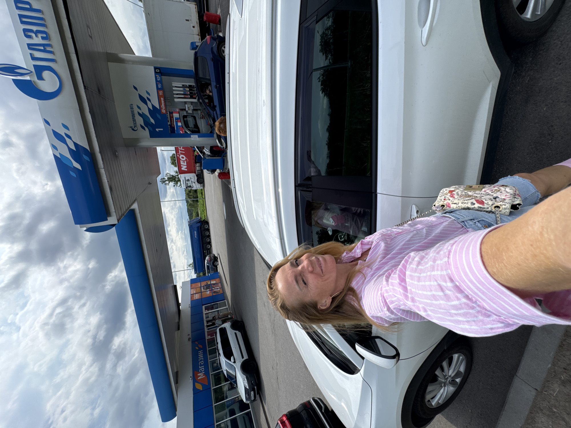 A woman in a pink and white striped shirt and denim shorts is taking a selfie at a gas station. She is standing next to a white SUV, with the pump area of the gas station behind her. The gas station has a large "GAZPROMNEFT" logo on it. There are other cars and trucks at the gas station, and a store with "Магазин" (Shop) written on it. The sky is cloudy.