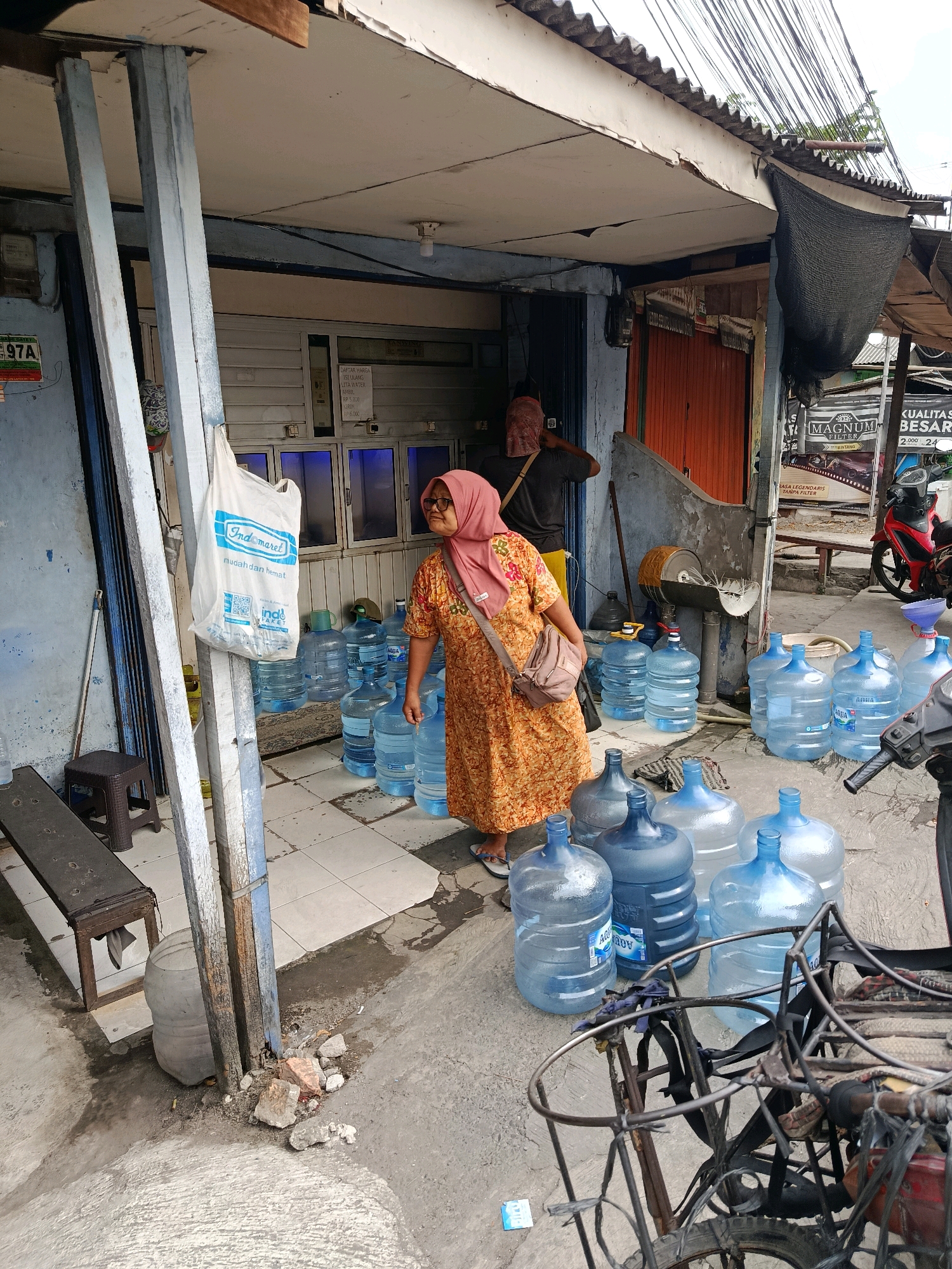 The image depicts an outdoor scene in Bekasi, Indonesia, likely a small roadside establishment selling bottled water. A woman wearing a pink headscarf and an orange floral dress stands among numerous large, clear plastic water jugs, some stacked on top of others. She appears to be looking to her left, with a light-colored shoulder bag slung across her chest. Behind her, a man in a dark t-shirt and what appears to be a red head covering is visible inside a structure with blue-tinted windows and metal shutters. The structure has a corrugated metal roof supported by wooden beams, and electrical wires are strung overhead. In the foreground, part of a metal bicycle rack is visible, with a portion of a red motorcycle to the right. A white plastic bag with the "Indomaret" logo hangs from a metal pole to the left. The ground is paved with square tiles and concrete, with some debris and puddles of water present. A wooden bench is situated to the left of the scene. The overall environment suggests a somewhat utilitarian, possibly informal commercial area.