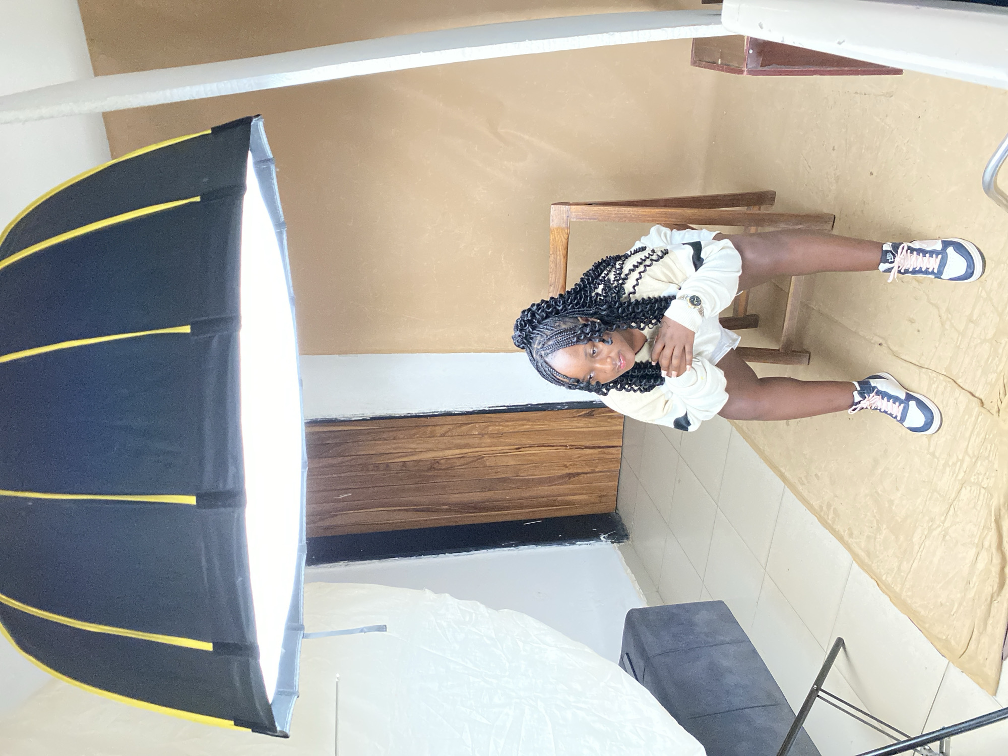 A medium-shot photograph depicts a young woman posing in what appears to be a photography studio. The studio is likely located in Jos, Nigeria, given the request specifications.
The woman is seated on a small wooden stool. She is wearing a light-colored, possibly off-white, outfit and sneakers that are predominantly white with dark blue accents. Her hair is styled in braids. She is facing the camera with her hands positioned near her face.
The studio setup includes a large softbox light modifier positioned to the left of the frame, providing illumination. There is also a white reflector placed in front of her. The background consists of a plain wall, and a wooden divider that is located behind her. The floor is covered in a beige rug, and tiles. Other studio equipment, like a bench and light stands are also partially visible.
