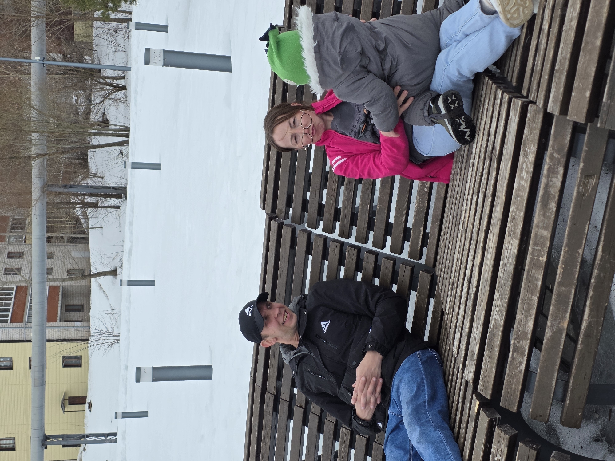 The image depicts a man and a girl sitting on a wooden bench in Votkinsk, Russia. Snow covers the ground and surrounding areas. The man is wearing a black Adidas jacket and a baseball cap. He is seated with his legs extended and his hands folded in his lap. The girl is wearing a pink jacket over a grey jacket, light blue pants, and a green hat. The background includes a row of white buildings with grey horizontal stripes and bare trees. There are also several short grey vertical poles sticking out of the snow.