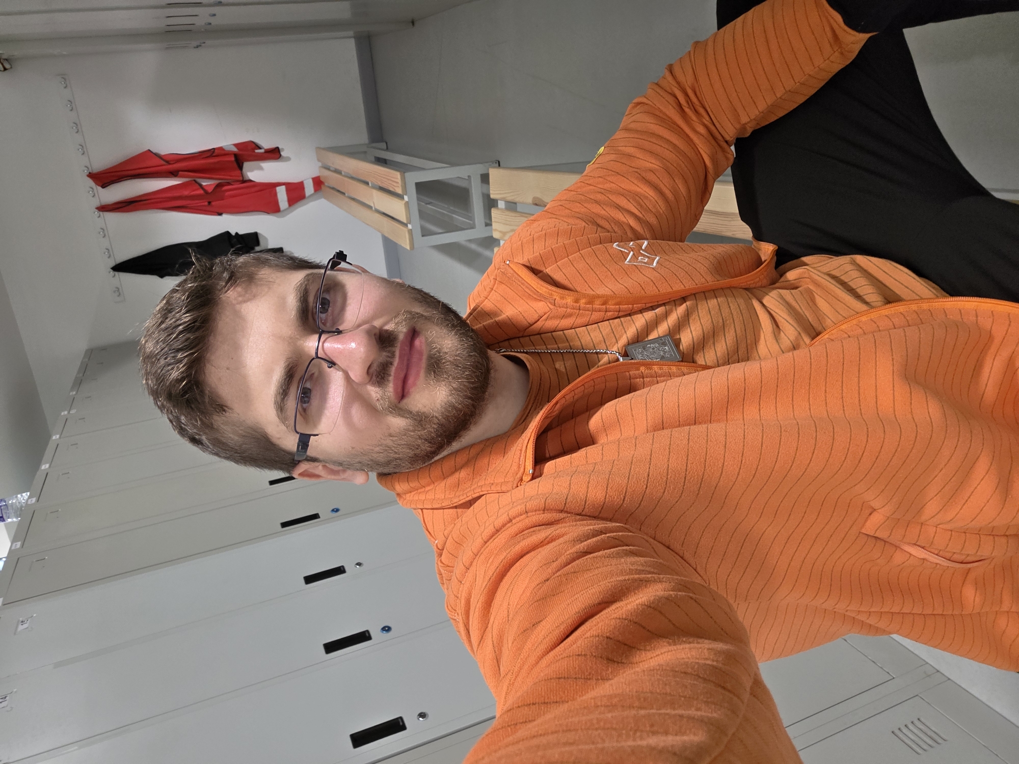 A man is taking a selfie in what appears to be a locker room. The man is wearing glasses and an orange striped hoodie. He is sitting with his legs bent, wearing black pants. Behind him are rows of gray lockers and a wooden bench. On the back wall, red and black garments are hanging on hooks. There is no information available to ascertain the location in Plovdiv, Bulgaria.