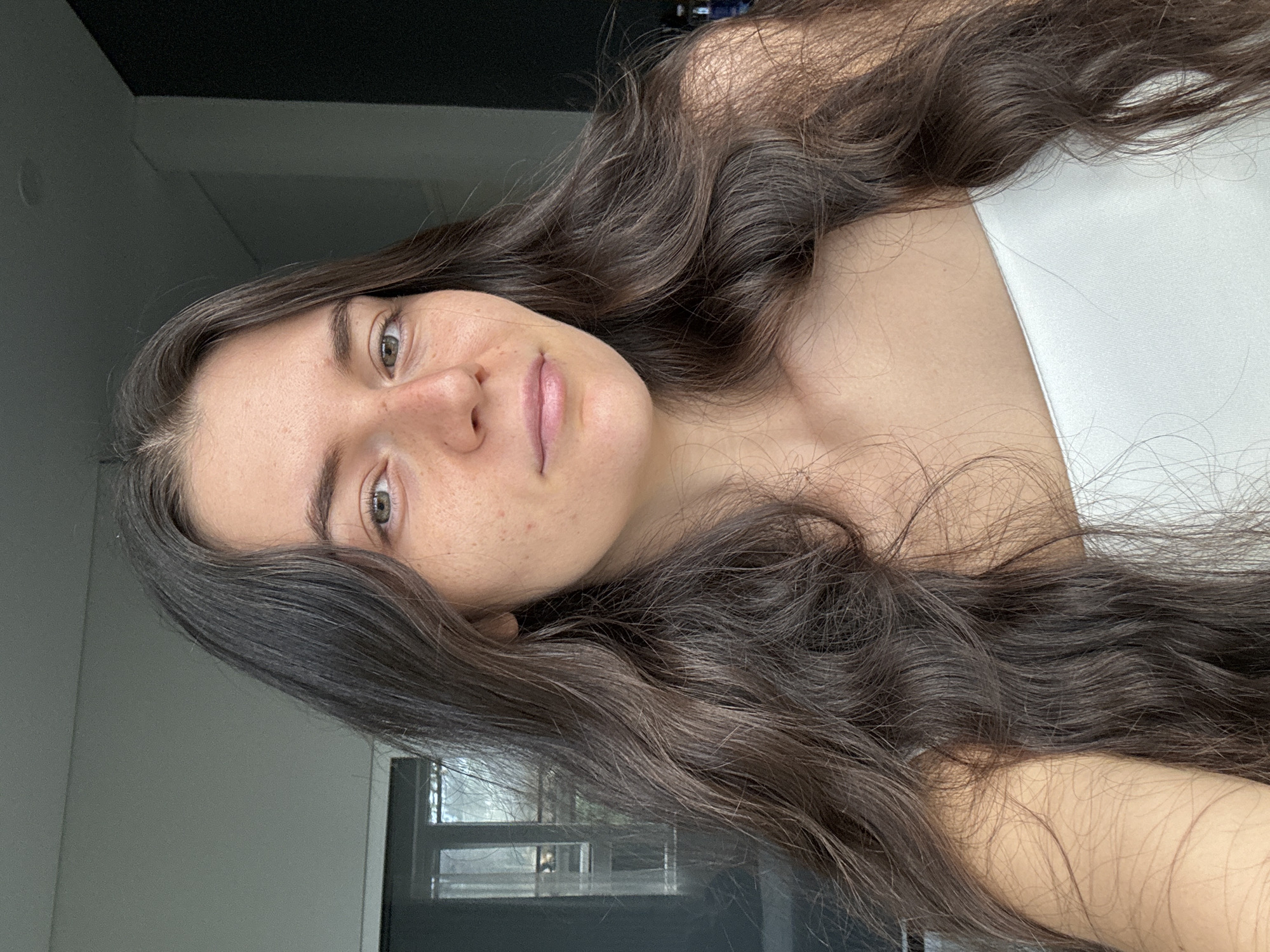 A young woman is pictured indoors with natural light filtering through a window. She has fair skin with some freckles and long, wavy, dark brown hair. She is wearing a white top. The background shows a neutral-colored wall, a window with a partial view of what appears to be foliage or trees outside. The location is not identifiable as Da Lat, Vietnam from this image alone.