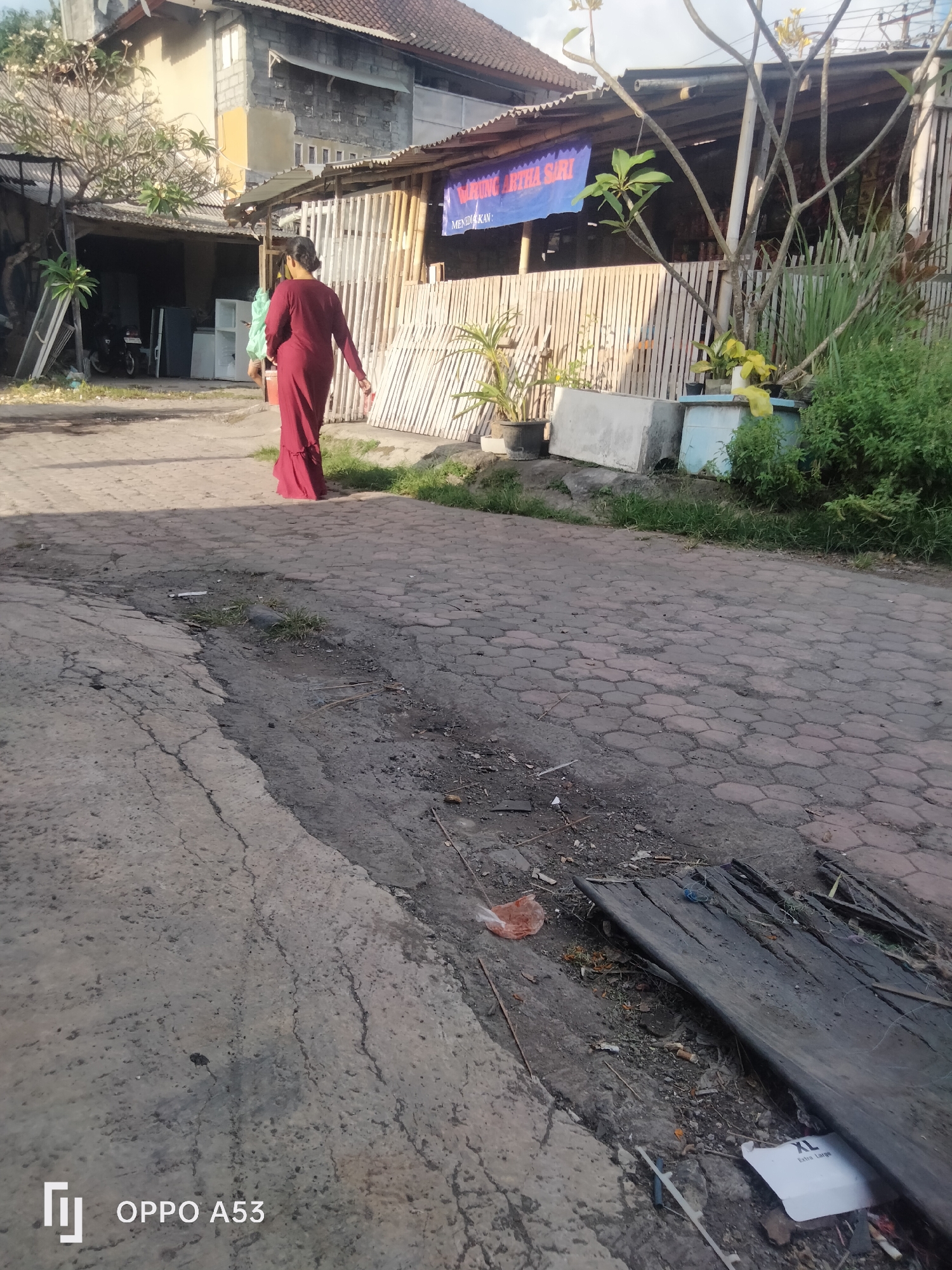 An outdoor, low-angle medium shot captures a woman walking away from the camera in a residential alley in Denpasar, Indonesia. The woman, dressed in a long, flowing, dark red dress, walks on a cracked concrete path. Behind her, the alley is lined with bamboo fencing and a long, low building with a corrugated metal roof. A blue banner with white text is visible on the building, along with a few potted plants and some discarded materials. The ground is a mix of cracked concrete, hexagonal paving stones, and debris. In the foreground, the cracked concrete path dominates the scene, with a shallow ditch running alongside it. The lighting suggests it is daytime, possibly late afternoon, with shadows cast by the building and vegetation. The overall impression is of a somewhat neglected urban environment.