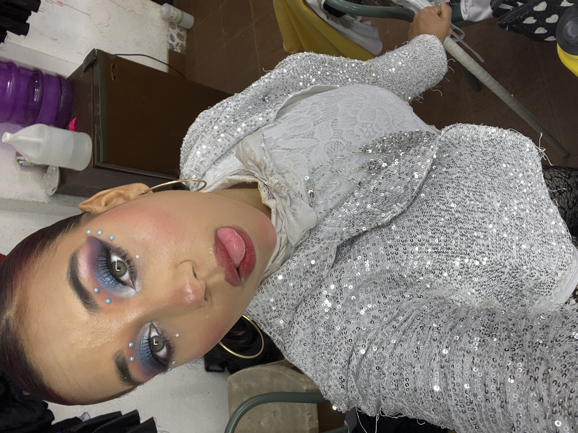 A young woman with dark hair pulled back is shown in a close-up, self-taken shot. She is wearing a silver sequined jacket over a white lace top and appears to be sticking her tongue out. Her makeup is elaborate, featuring blue eyeshadow with small pearl embellishments around her eyes and blush on her cheeks. The background includes a dark brown cabinet, a large purple water jug, and other assorted items, suggesting an indoor, possibly backstage or dressing room setting. The lighting is somewhat dim and uneven, casting shadows.