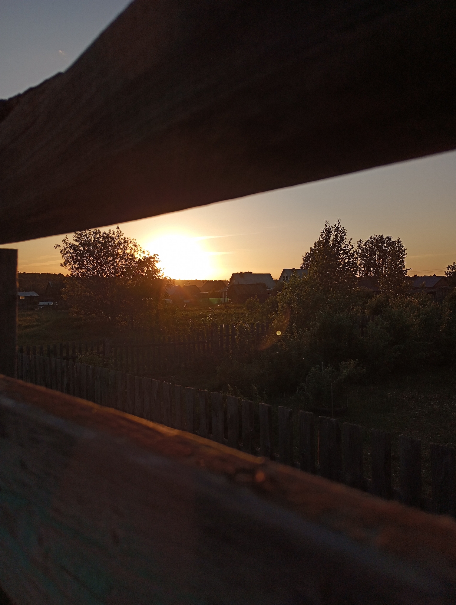 The image shows a view through a wooden structure, possibly a fence or a partially constructed building, towards a landscape with trees, houses, and a setting sun. The location is Буренка, Russia. The sun is positioned near the horizon, casting a warm glow over the scene. A wooden fence extends across the middle ground, separating the foreground from the background. Beyond the fence, there are trees, bushes, and several houses, suggesting a rural or suburban setting. The overall scene suggests a quiet, peaceful moment in the late afternoon or early evening.