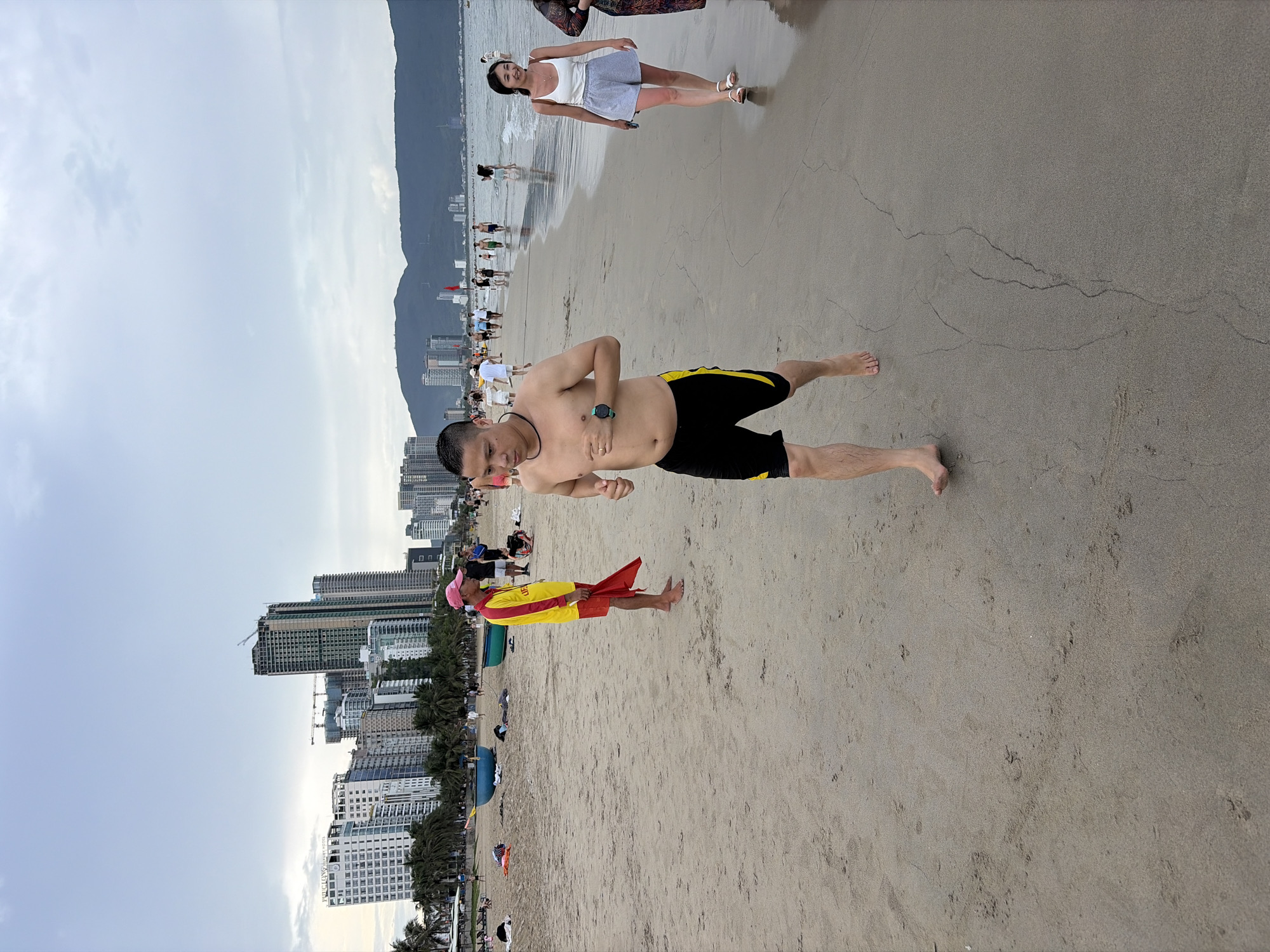 The image depicts a busy beach scene in Đà Nẵng, Vietnam, with the city's skyline visible in the background. In the foreground, a shirtless man with short dark hair is running on the sand, wearing black shorts with yellow trim and a black necklace. To his left and further back, a lifeguard in a yellow and red uniform stands with a red flag. Several other beachgoers are scattered across the sand, some walking along the shoreline. The beach itself is a light brown sand, with gentle waves visible in the distance. The sky is overcast with scattered clouds. In the background, numerous high-rise buildings line the coast, and mountains can be seen in the far distance.