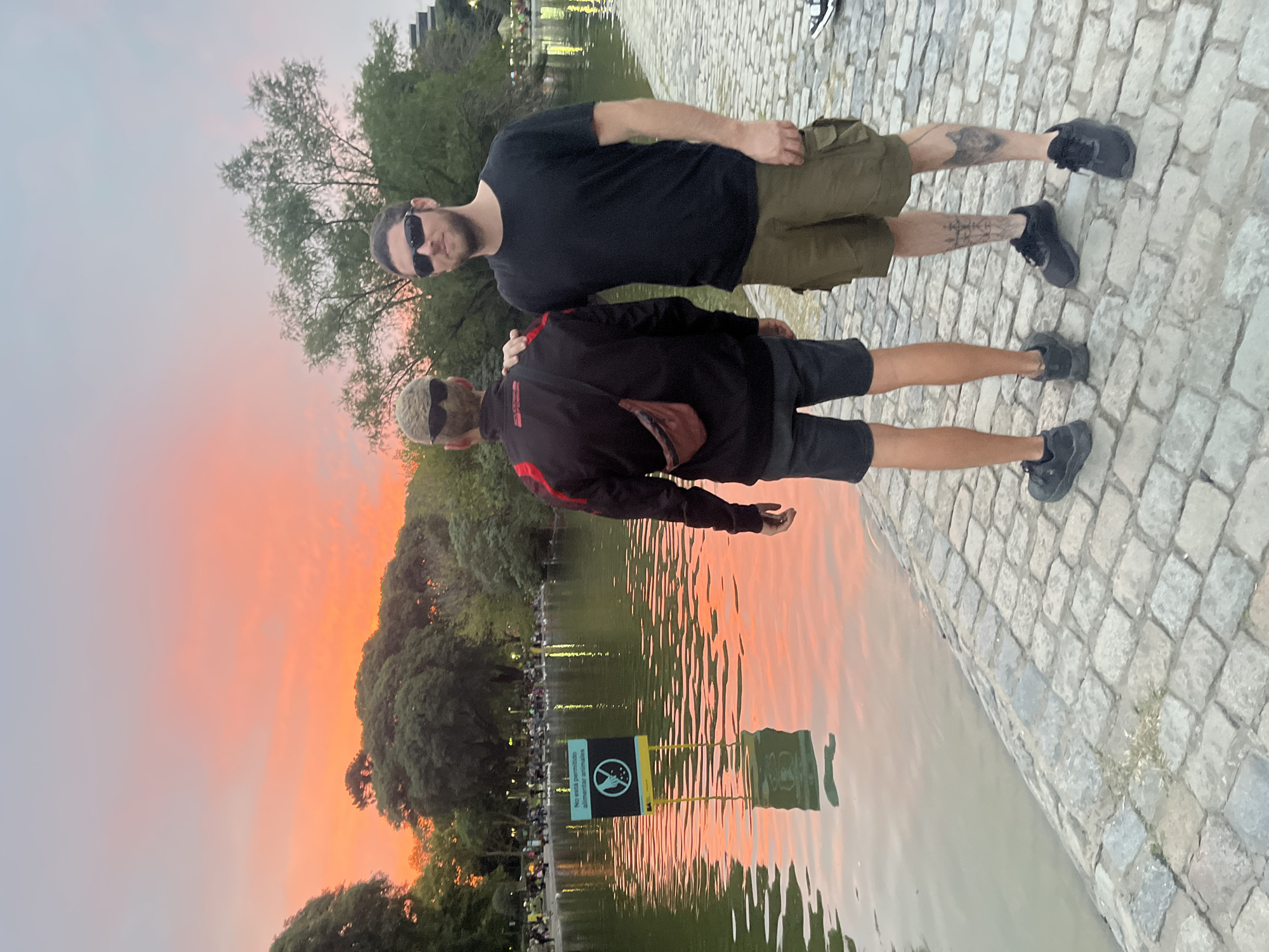 Two men stand side-by-side on a cobblestone path next to a body of water at dusk. The man on the left wears sunglasses, a black t-shirt, and olive-green shorts. Tattoos are visible on his right leg. The man on the right wears sunglasses, a black jacket with red accents, black shorts, and a small brown bag. A sign prohibiting feeding animals is visible in the water, reflecting the orange-hued sky. The location is the Paseo El Rosedal in the Bosques de Palermo, Buenos Aires, Argentina.