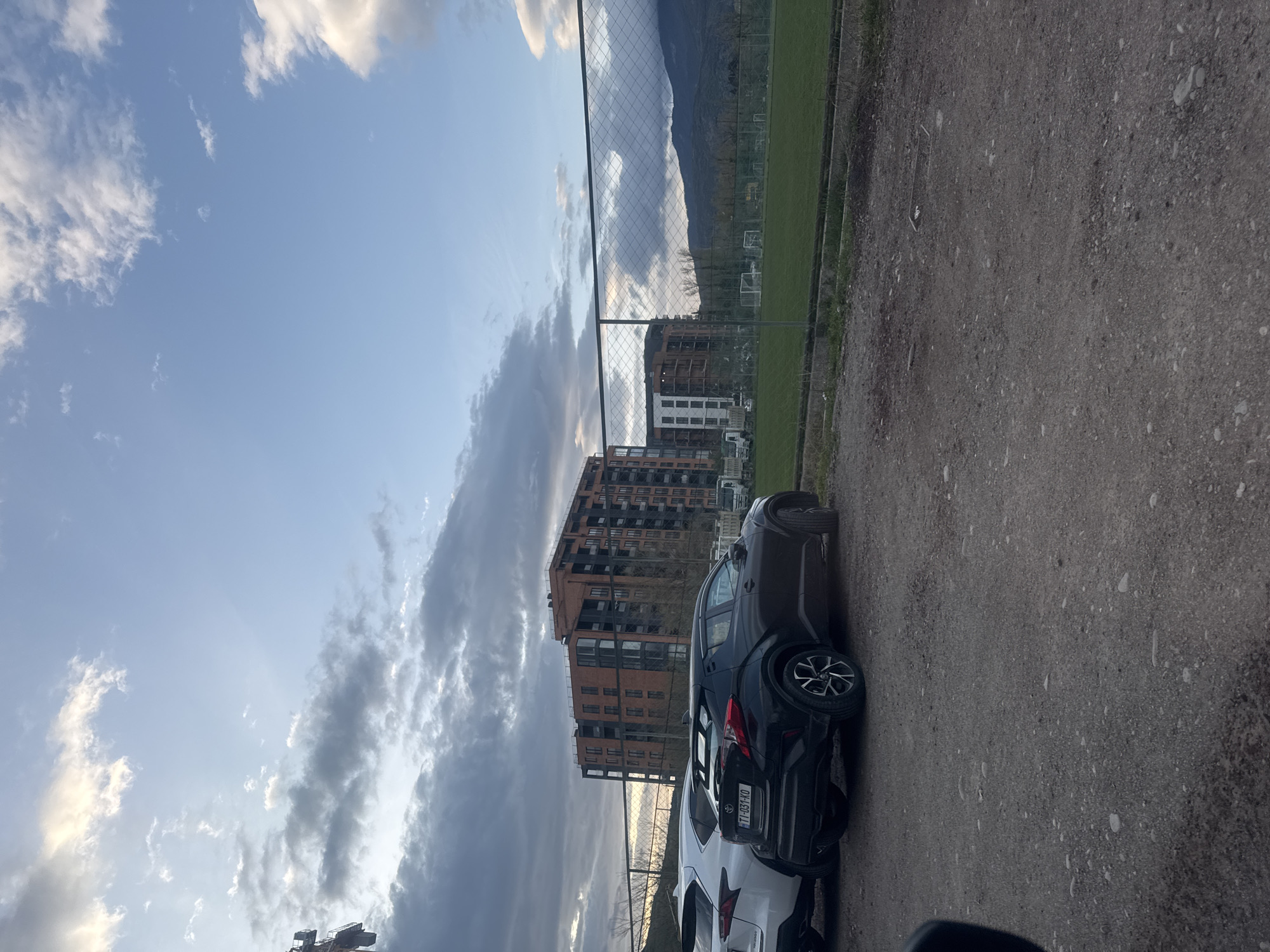 The image depicts a view in Tbilisi, Georgia. The scene shows a row of parked cars alongside a gravel area adjacent to a soccer field. A chain-link fence separates the parking area from the field. Behind the field, there are apartment buildings and a mountain range in the distance. The sky is partially cloudy with a mix of blue sky and grey clouds. The lighting suggests either early morning or late afternoon. One of the cars has a Georgian license plate.