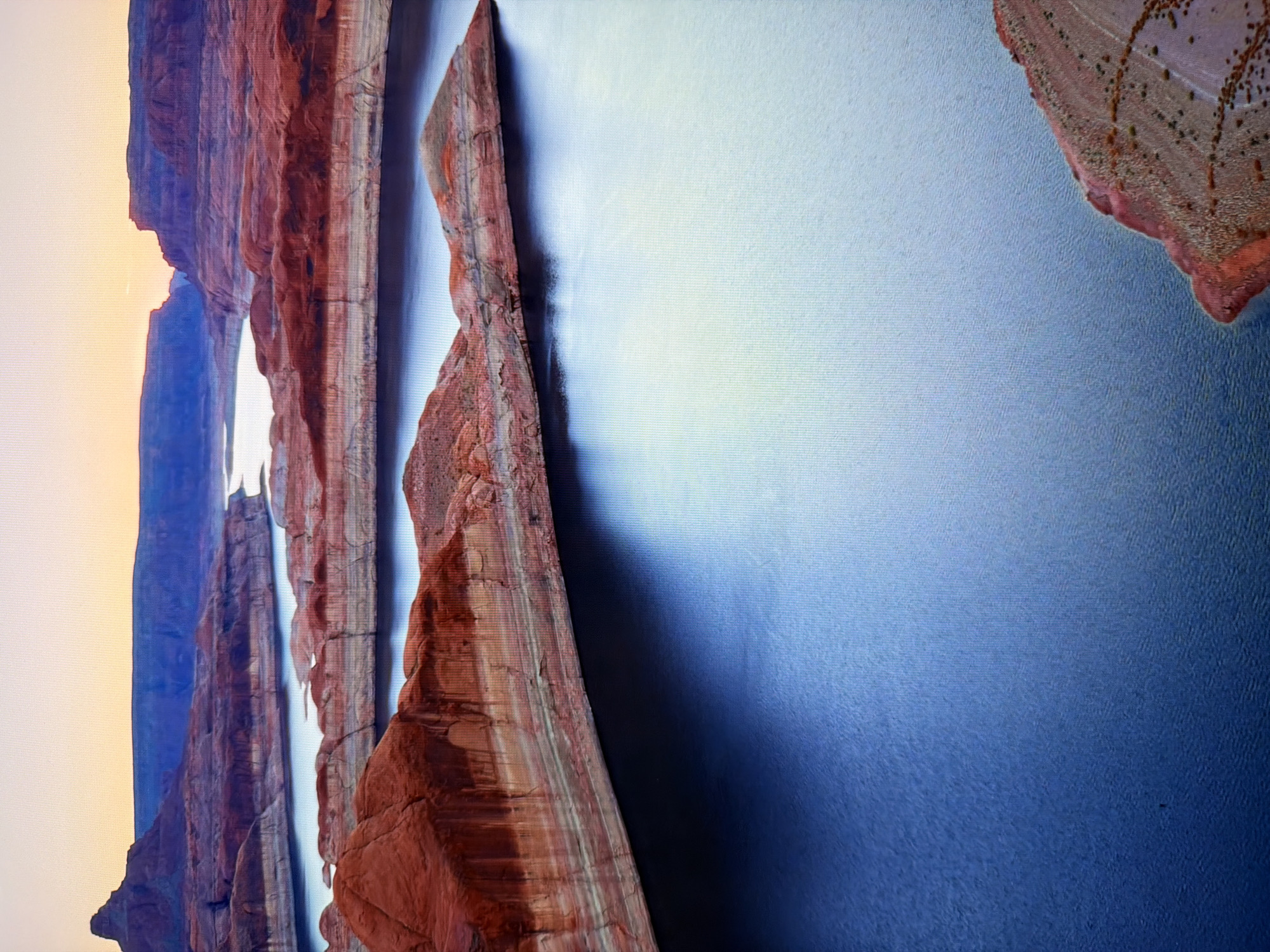 This is a vertical, aerial drone shot of Lake Powell, Arizona, at sunrise. The sky is a gradient of pale yellow and orange at the horizon, transitioning to a soft blue. Layered, red and white sandstone mesas with sharp edges and flat tops dominate the middle ground, with water filling the canyons between them. The water is a deep, dark blue in the foreground, becoming lighter and more reflective towards the background where it merges with the sky. The foreground features a reddish-brown, textured rock formation jutting into the lake from the lower right corner. The image is clear and sharp, with no discernible people or man-made structures visible, indicating a natural landscape scene.