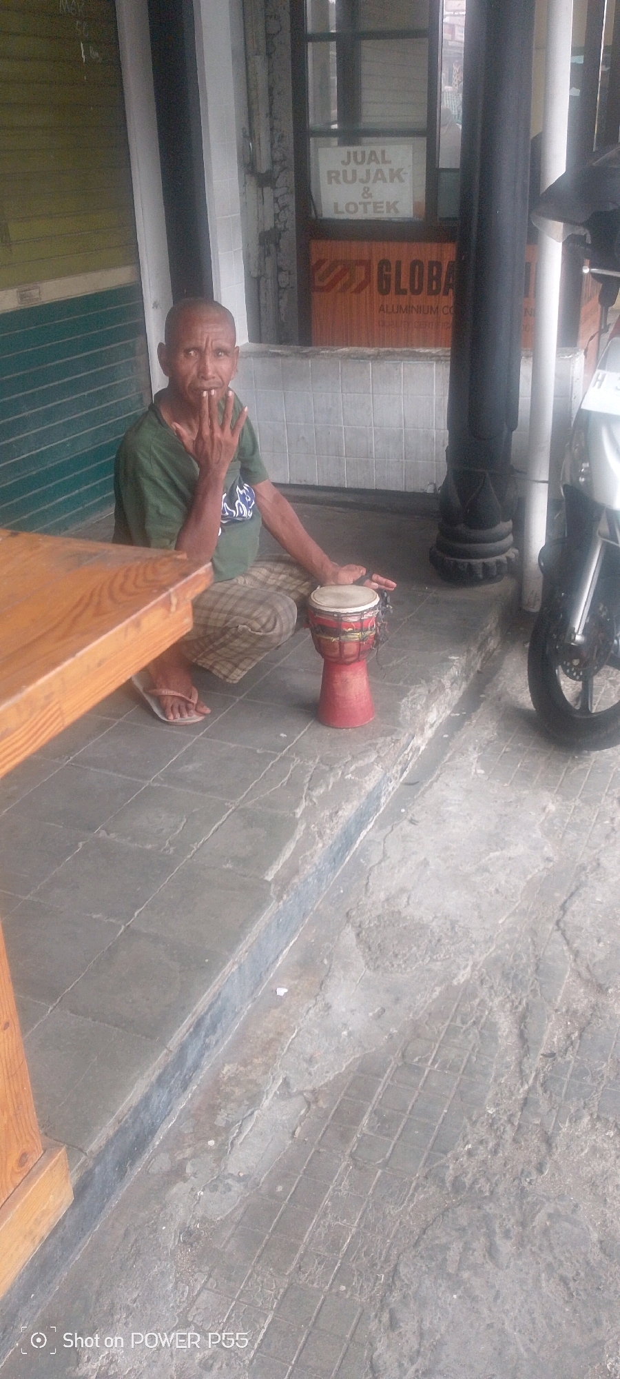 An older East Asian man wearing a green t-shirt and plaid pants kneels on a gray tiled ground next to a wooden table and a small red drum. The man is looking at the camera with his right hand raised near his mouth. A motorcycle is parked to the right of the scene. Behind the man, there is a building with a window displaying signs for "JUAL RUJAK & LOTek" and "GLOBAL ALUMINIUM CO." The ground is a mixture of tiles and rough concrete.