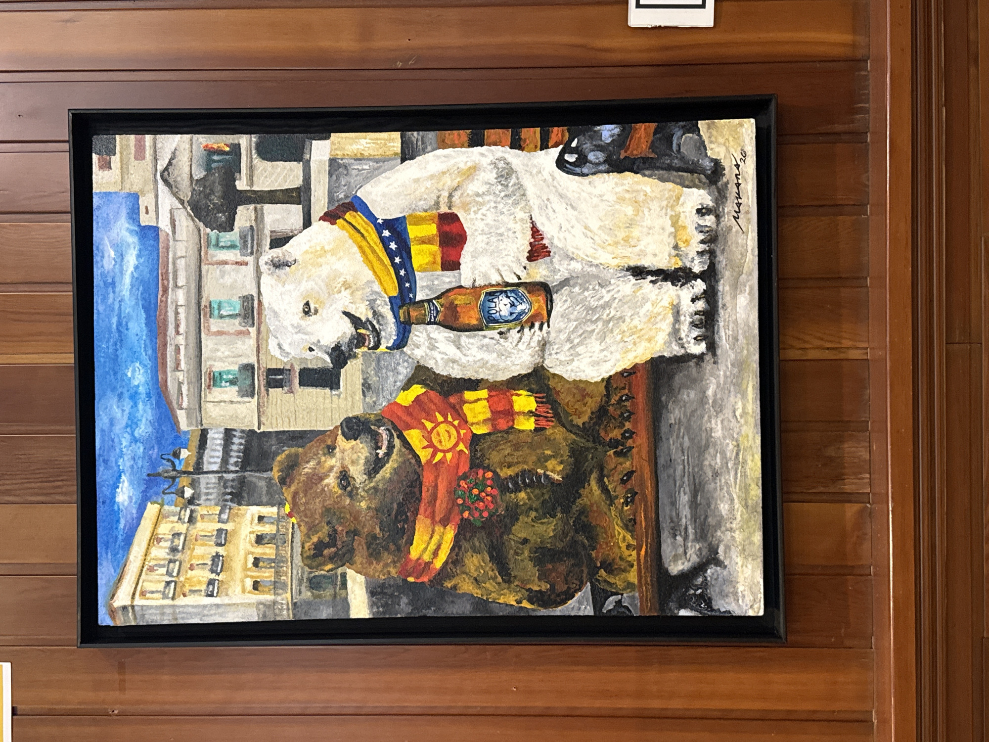 This is a painting of two anthropomorphic bears sitting on a park bench in front of buildings that appear to be in a European city, possibly Spain. The bear on the left is brown and wearing a scarf with a sun emblem. The bear on the right is white and holding a bottle of Polar brand beer. Both bears are smiling. The background shows buildings with balconies and windows, and a lamppost. The painting is framed in black and hangs on a wood-paneled wall. The signature on the painting reads "Morans '20". The location is not specified within the image itself.