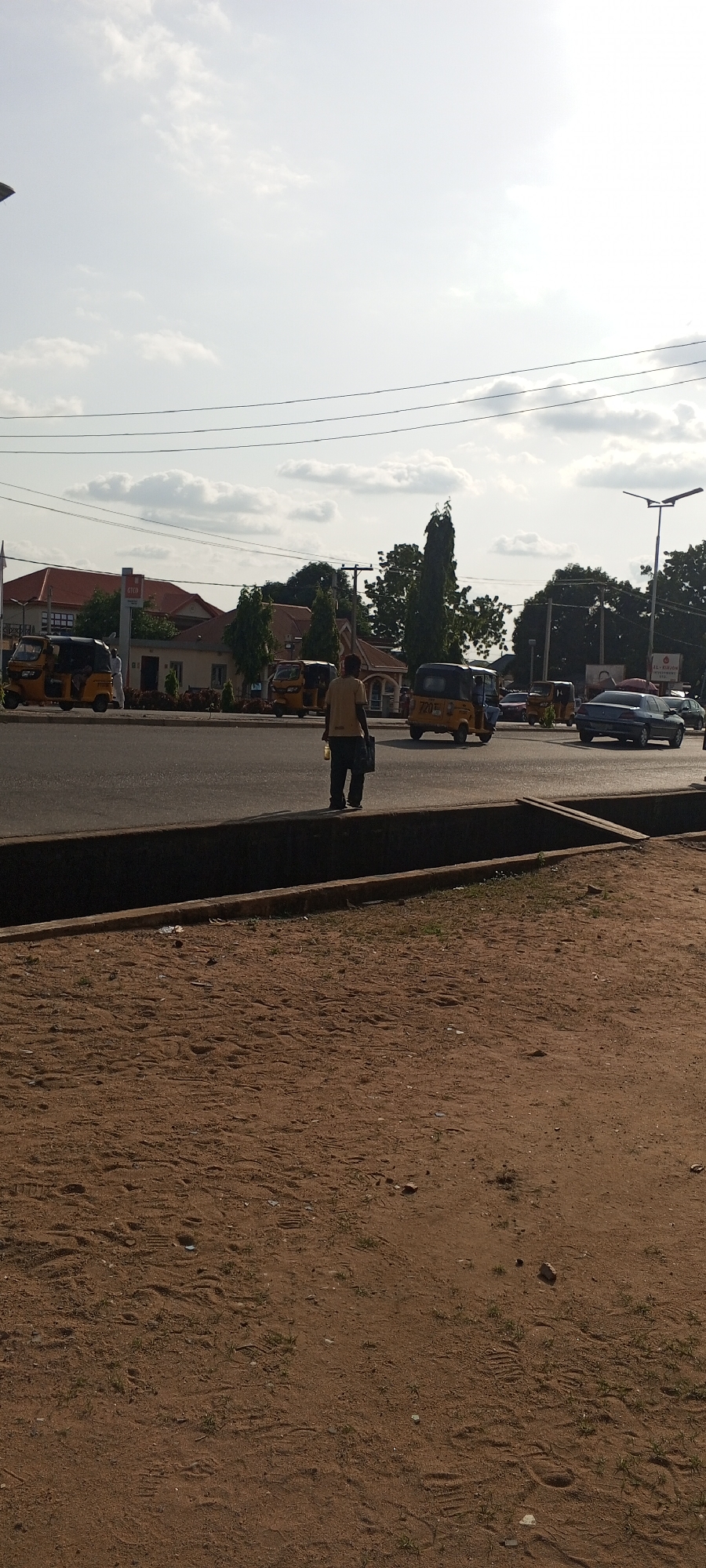 This image captures a street scene in Jalingo, Nigeria, under a bright, partly cloudy sky, suggesting it is daytime. The foreground features a sandy, disturbed area with visible footprints and sparse vegetation, bordering a concrete drainage channel.
The main subject is a person standing on the edge of the road, facing away from the camera. They are wearing a light-colored t-shirt and dark trousers, and holding a dark bag or briefcase in their right hand. Their posture appears casual, possibly waiting or observing.
The background reveals a busy street with multiple vehicles. Several yellow, three-wheeled auto-rickshaws, commonly known as "Keke Napep" or "Marwa," are visible, some in motion and others parked. A blue sedan is also present on the road. Beyond the street, buildings with red-tiled roofs and some modern structures are discernible, interspersed with tall, slender trees and electrical wires strung across the scene. A street lamp is visible on the right side of the image. The overall atmosphere suggests an urban environment during a typical day, with the activity centered around traffic and pedestrians on a main road. No discernible text is visible in the image.