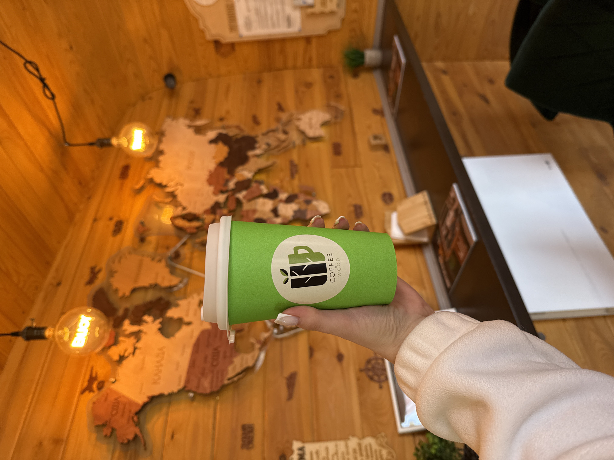 A hand holds a green coffee cup with a white lid and a logo that says "Coffee & Wood." The background is a wooden wall decorated with a map of the world. There are also light bulbs and other decorative items on the wall. The location appears to be inside a coffee shop. The city and state are unknown.