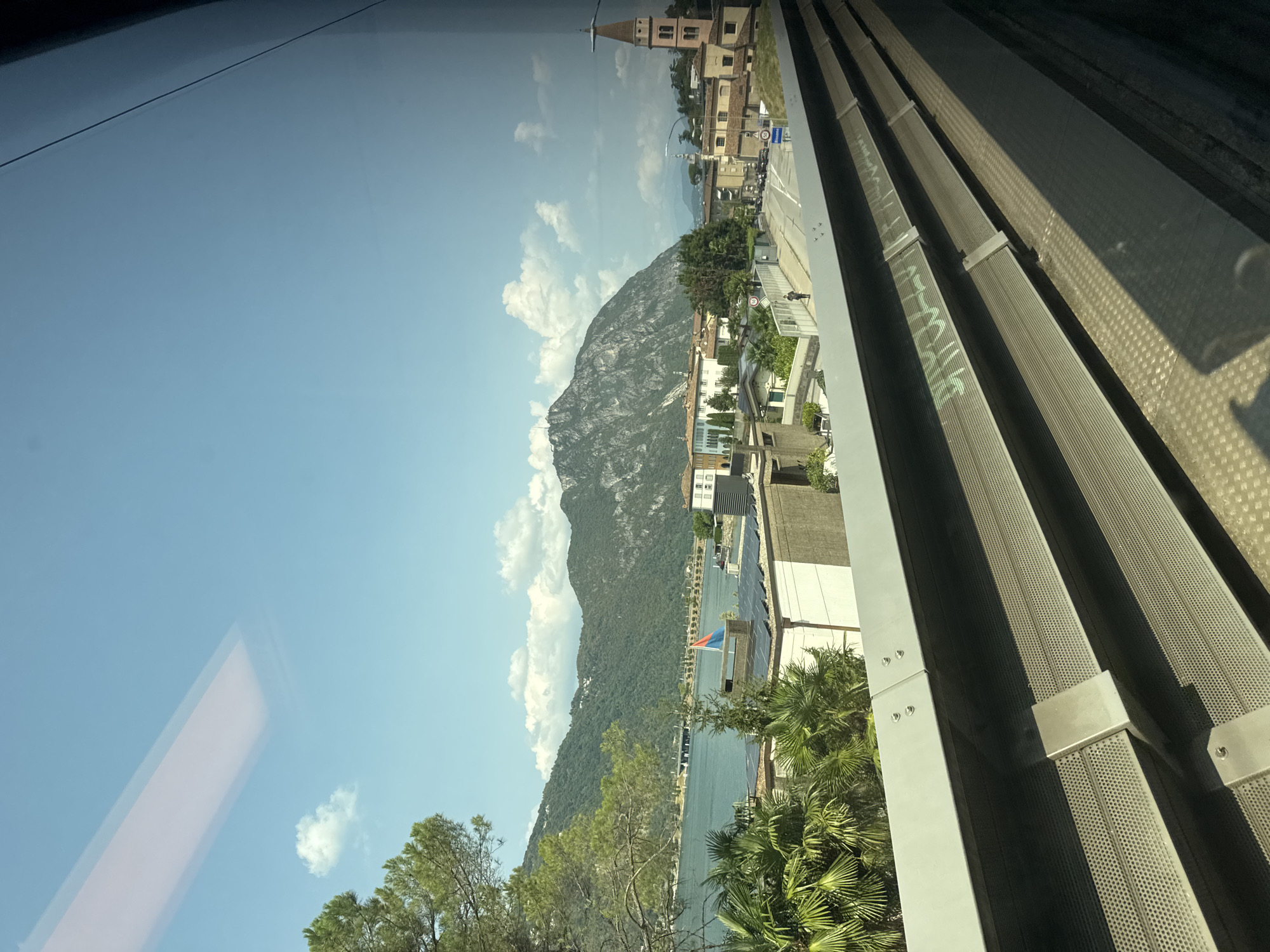 A horizontal, eye-level, slightly tilted view depicts the town of Bissone, Switzerland, from the perspective of a train. In the foreground, the train's interior structure is visible, consisting of metal grating with a textured, perforated surface. The middle ground showcases a cityscape with buildings featuring white facades and tiled roofs, interspersed with lush green palm trees and other vegetation. A body of water, likely a lake, stretches across the left side of the frame, bordered by a bridge and rolling green hills. Majestic mountains dominate the background, their peaks partially obscured by the bright, blue sky dotted with scattered white clouds. A single person is visible walking on a paved path in the mid-ground. The lighting suggests a sunny day.
