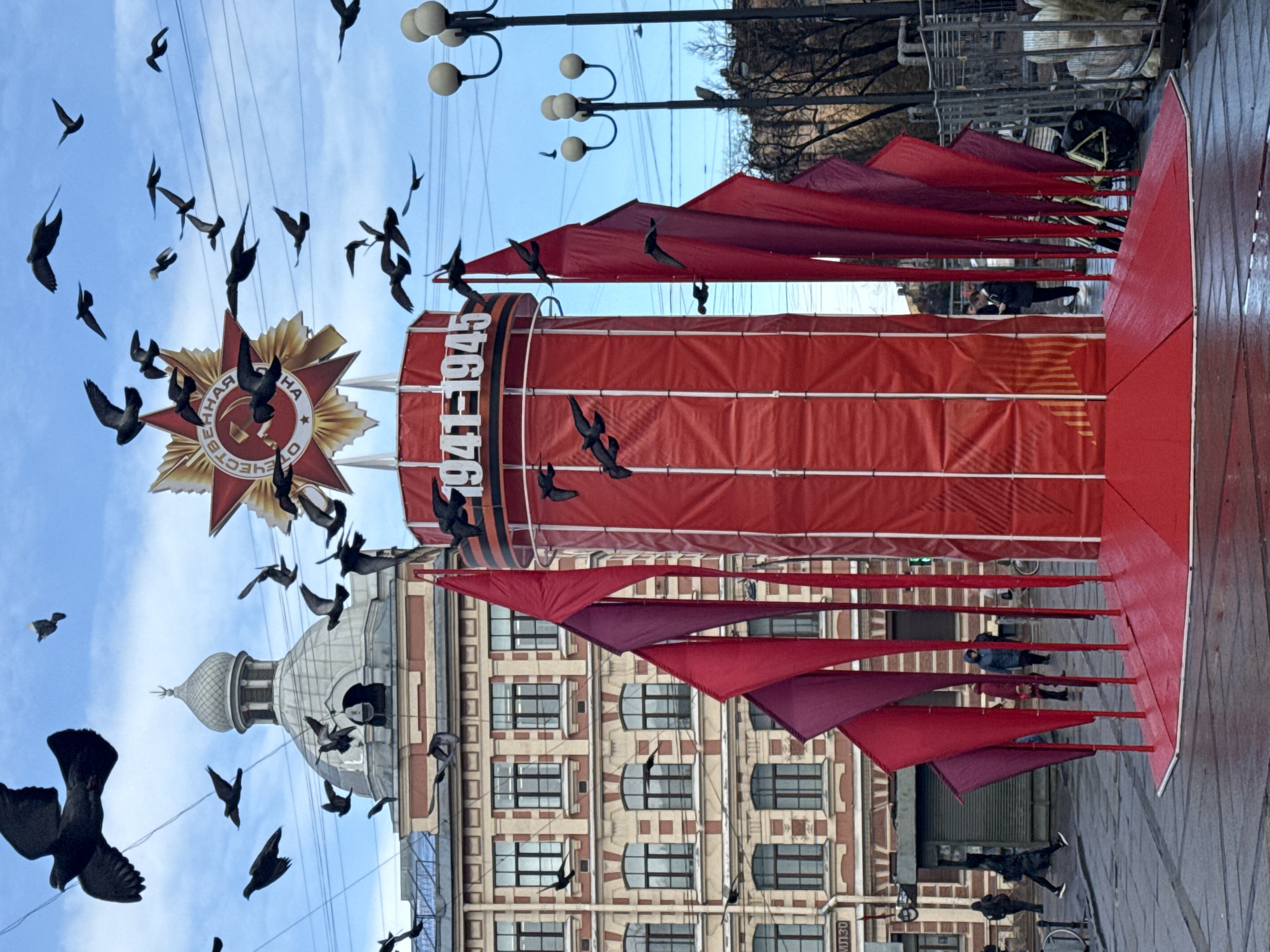The image captures a scene in Saint Petersburg, Russia, featuring a large, red, cylindrical structure adorned with Soviet-era symbols. At the top of the structure is a prominent red star with the inscription "ОТЕЧЕСТВЕННАЯ ВОЙНА," which translates to "Patriotic War," and the years "1941-1945" marked below. Red banners extend outwards from the structure. Numerous birds are in flight around the monument and in the sky. A building with a light-colored dome and a facade with multiple windows can be seen in the background. Street lights are visible overhead, and people are walking along the wet pavement at the base of the structure.
