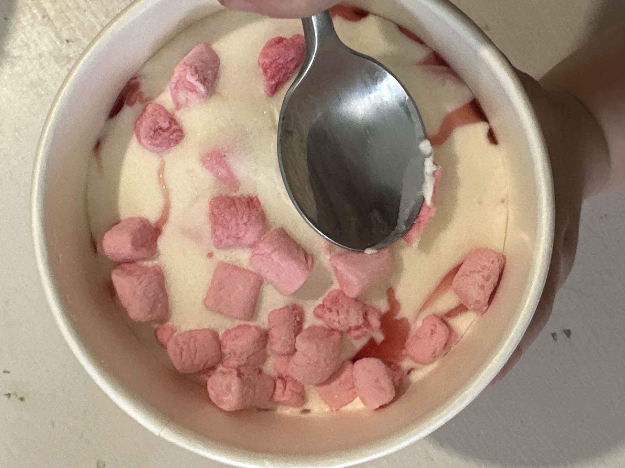 A top-down shot reveals a white paper pint container filled with light yellow ice cream interspersed with pink, cube-shaped marshmallows. A silver spoon is positioned in the upper center of the container, seemingly in the act of scooping the ice cream. Part of a hand or arm is visible on the right side of the frame, holding the container. The background is a light beige or white surface with a few scattered dark specks. There is no information about the city of Votkinsk, Russia, in the image.