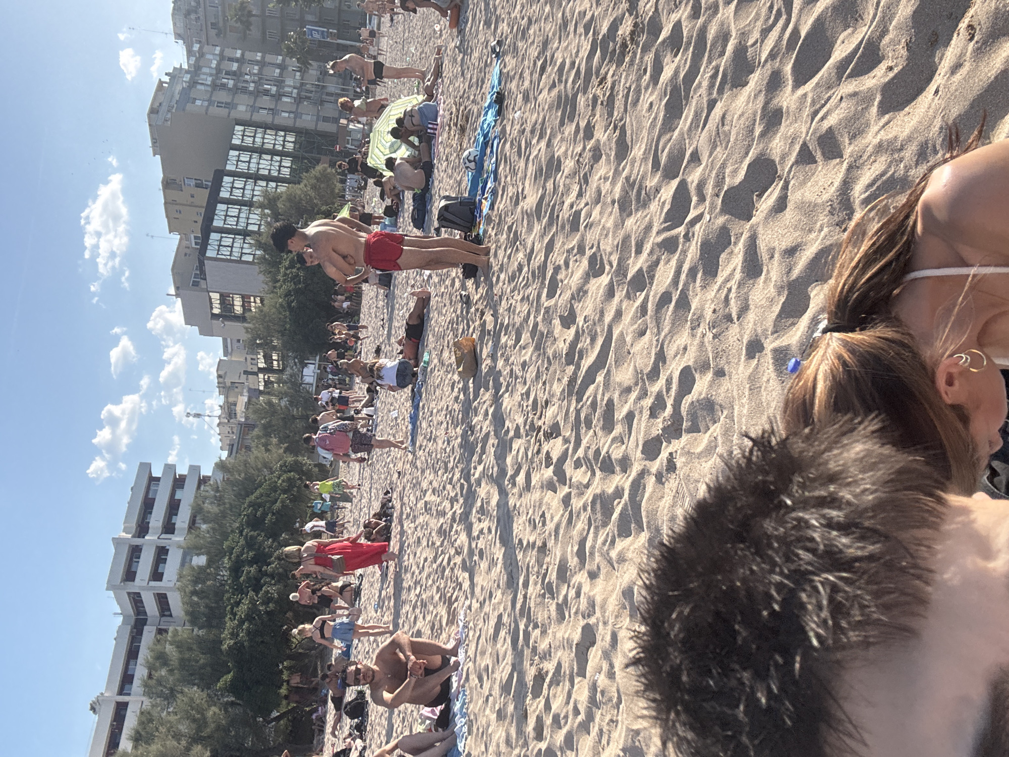 This is a sunlit, crowded beach scene, rotated 90 degrees counter-clockwise. The image, if corrected, would show a wide expanse of sandy beach bustling with people under a clear blue sky with scattered white clouds.
The primary subjects are numerous beachgoers, engaged in various leisure activities. Many are standing, walking, sitting, or lying on towels and blankets across the sand. In the mid-ground, a shirtless man in red swim shorts stands prominently, seemingly talking or interacting with another person whose back is partially visible. Near them, two individuals appear to be embracing or standing very close. Further into the background, a blue beach towel with a soccer ball and a black backpack lies on the sand next to a green and yellow striped beach umbrella, under which several people are seated. Other individuals are scattered throughout the beach, some sunbathing, others strolling, and many interacting in small groups.
The background features multi-story buildings and dense green trees, suggesting an urban or resort coastal location. The buildings vary in architectural style, with some having large reflective windows. The foreground is dominated by the textured sand, showing ripples and footprints. In the immediate foreground, the back of a person's head with dark, spiky hair is visible, along with the shoulder and part of the head of another person with long brown hair tied back, who is wearing a white swimsuit top and golden hoop earrings.
The activity taking place is typical of a busy beach day, with people enjoying the sun and sea. The bright sunlight and clear sky indicate good weather. There is no visible text in the image. The time of day appears to be daytime, likely late morning or afternoon, given the shadows. The location cues point to a developed coastal area, but the exact geographical location remains unknown.