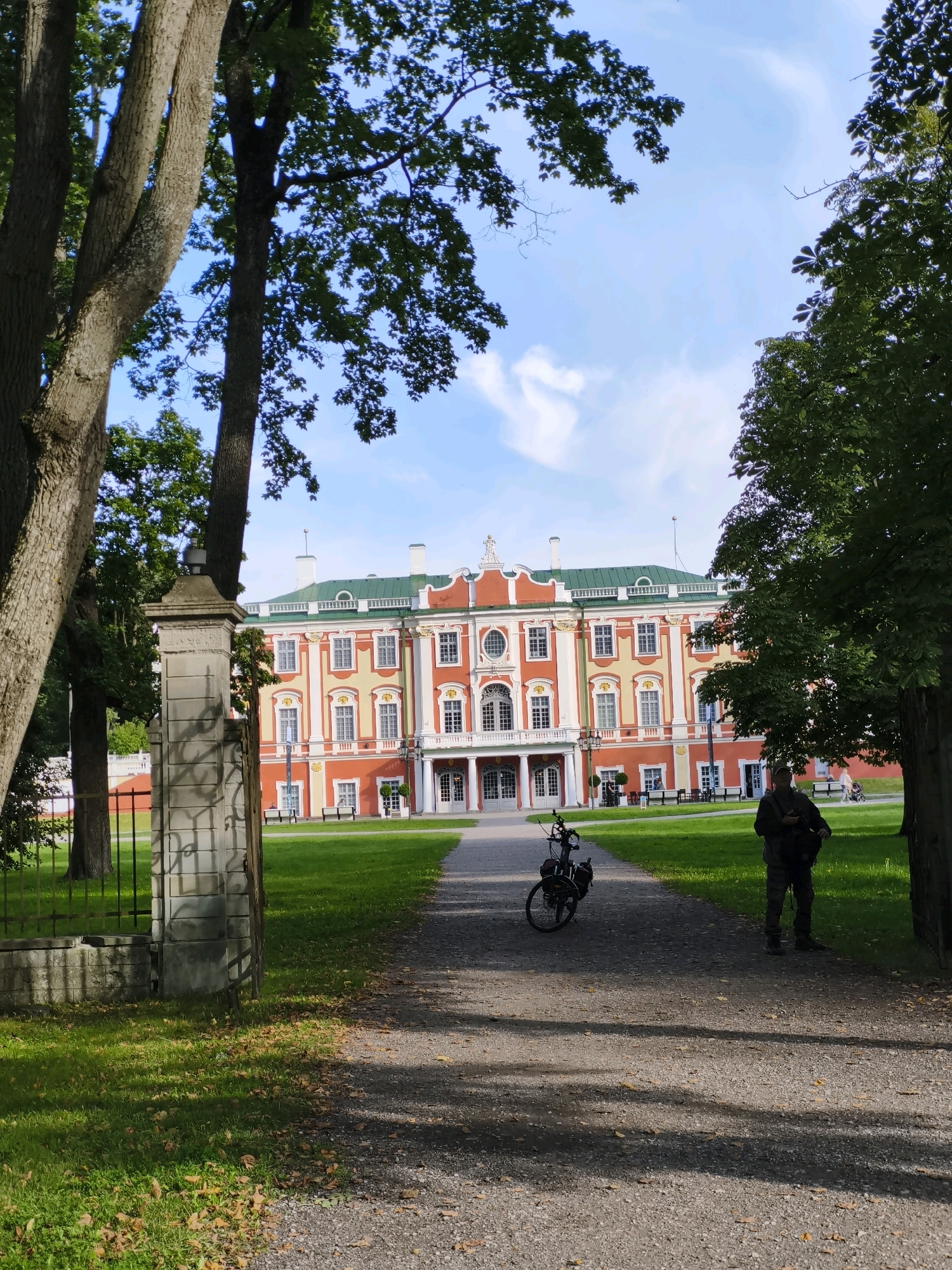 The image depicts the Kadriorg Palace in Tallinn, Estonia, a Baroque palace and a museum. In the foreground, a gravel path leads towards the palace. On the path, a bicycle is parked on the left, and a person is standing on the right, facing the palace. The person is wearing a dark jacket, pants, and a backpack. The palace is a large, ornate building with a red and yellow facade and a green roof. It is surrounded by a manicured lawn and tall trees. The sky is blue with scattered white clouds.