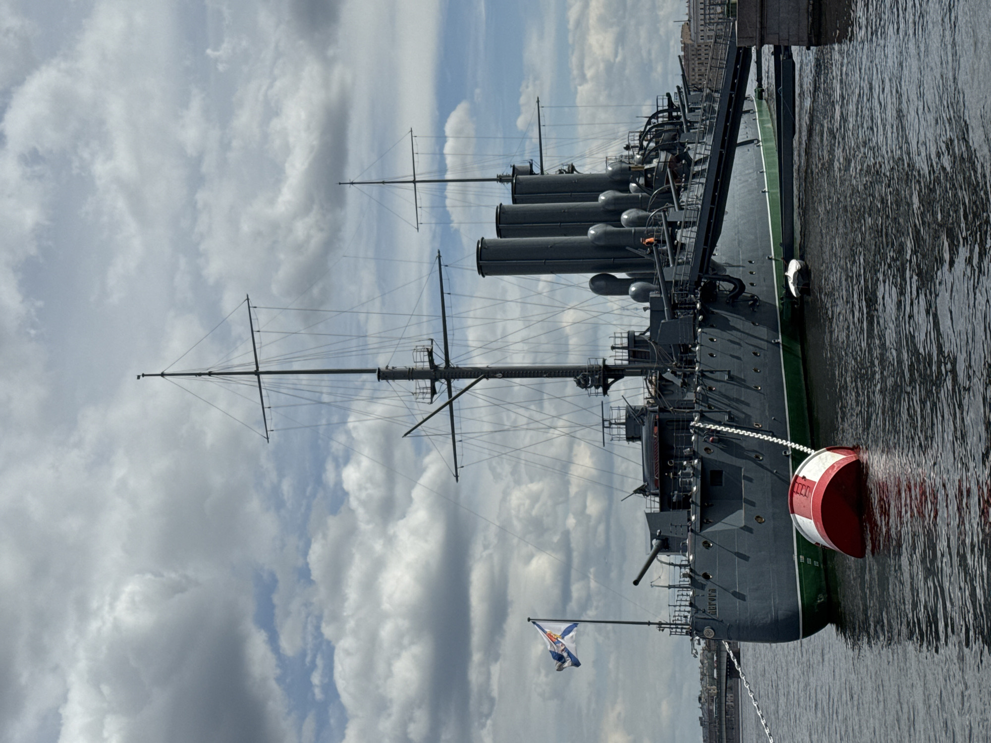 This image captures the historic cruiser Aurora docked in what appears to be Saint Petersburg, Russia, based on context and the ship's identity. The scene is set during the daytime under a partially cloudy sky.
The prominent subject is the battleship, painted in a dark grey hull with a distinct light green stripe along its waterline. Its large structure features multiple masts with intricate rigging, several prominent smokestacks (funnels), and visible gun turrets along its deck. The Cyrillic name "АВРОРА" (Aurora) is clearly visible on the ship's bow. At the stern, a Russian Naval Ensign (St. Andrew's flag) is flying, a white flag with a diagonal blue cross.
The ship is moored in dark, rippled water, which reflects the overcast sky and parts of the ship. A large, red and white striped buoy is floating next to the ship's stern, connected by a chain. In the distant background, faint architectural details of buildings on the opposite bank are visible.
The sky is largely covered with white and grey clouds, but enough light penetrates to illuminate the scene clearly, suggesting a bright, yet overcast, day. The overall impression is one of a preserved historical vessel, maintaining its imposing presence. No people are explicitly visible in the frame.