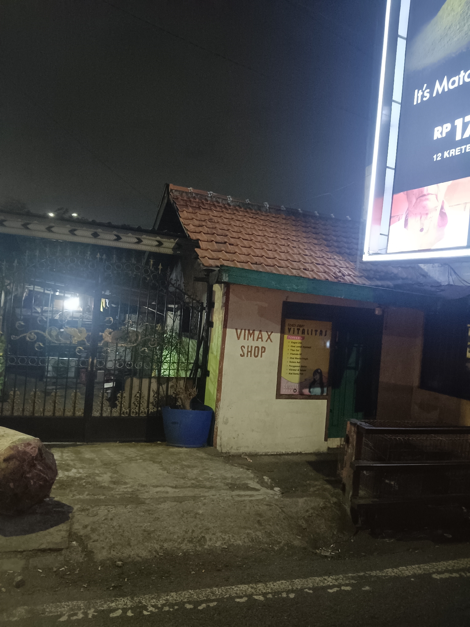 A nighttime outdoor shot shows a store called "VIMAX SHOP" in Medaeng, Indonesia. The store has a light-colored facade with a red-tiled roof. A large sign on the right side of the building displays "VITALITAS" and lists several products, including "Viagra USA," "Obat Lemak," "Titan Gel," "Vitamins AF," "Obat Kopi Pria," "Pangemuk Badan," and "Vibrator & Yakini." The entrance to the store is a dark doorway. In front of the store is a black wrought-iron gate. A blue planter with a dead plant is situated near the gate. The ground is paved with concrete. A large, rough-textured rock is visible on the left side of the image. A large illuminated billboard is also present on the right side of the image, partially obscured by the building.