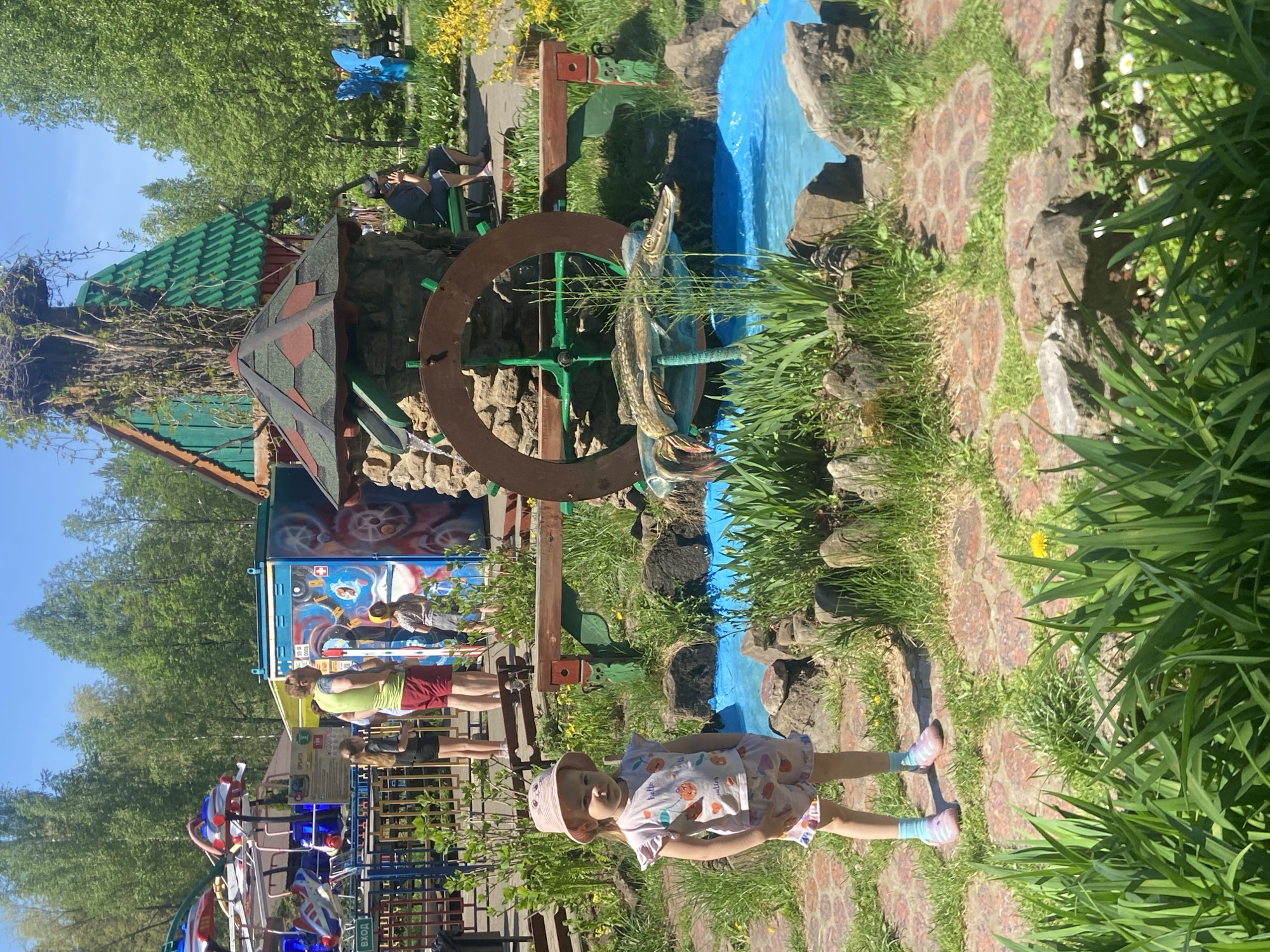 A young child in a light-colored dress and matching hat stands on a brick-paved path in an outdoor setting. Behind the child is a decorative water wheel and a faux stream with a fish sculpture. Adjacent to this is a small, ornate building with a stone base and a green tiled roof. In the background, there is a blurred amusement park area with rides and people. The scene appears to be taken in a park or recreational area in Chaikovsky, Russia, judging by the presence of specific amusement park features and architectural details. The environment includes trees, grass, and other foliage.