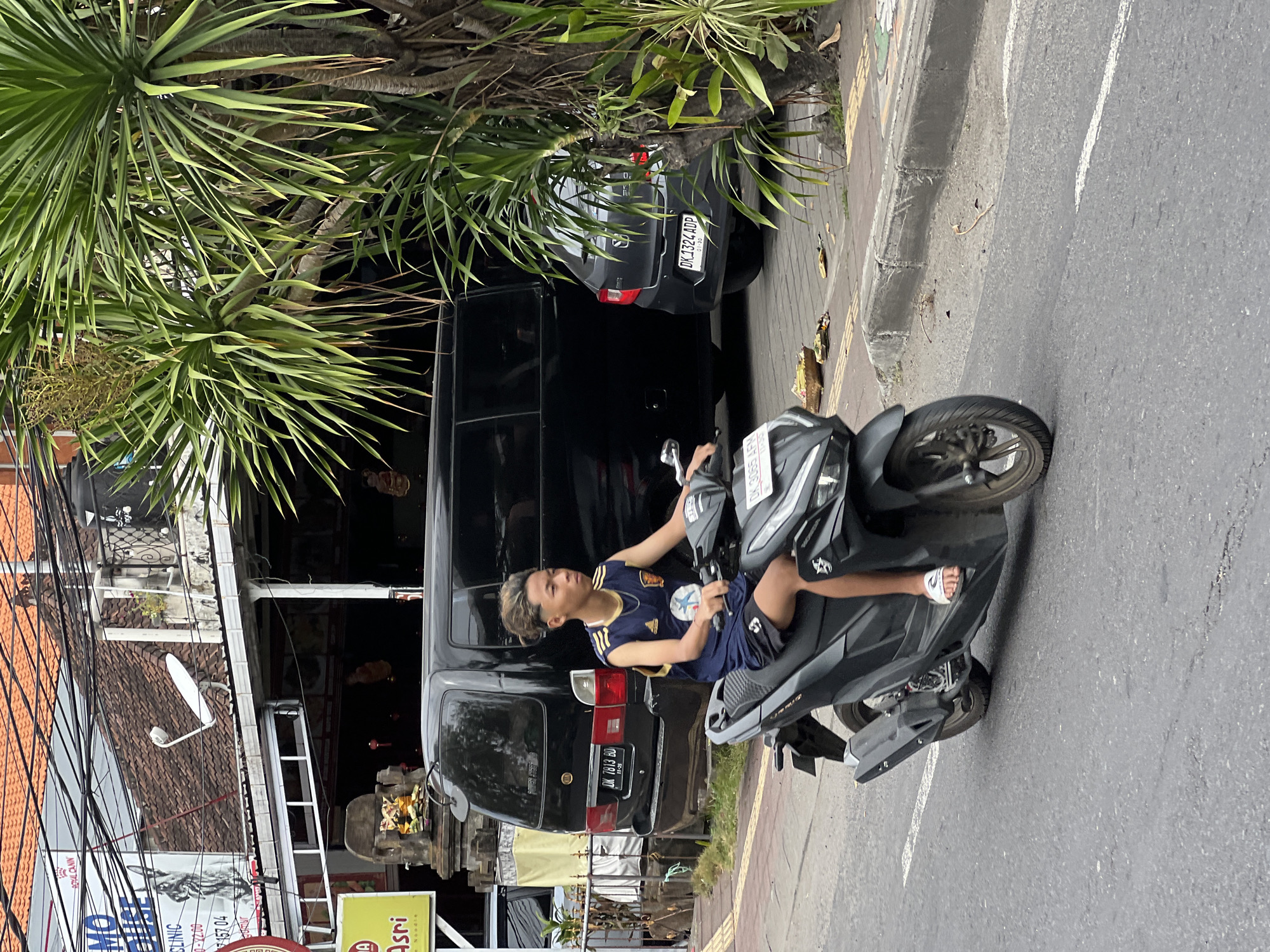 A young male with blonde-streaked, styled hair rides a black and gray scooter down a paved street in Denpasar, Indonesia. He wears a dark blue sleeveless jersey with yellow trim, a white athletic logo on the front, and black shorts. The scooter has a license plate that reads "DK 3065 AFN." In the background, a black van with license plate "DK 7813 BD" is parked, and to its right, a dark gray car with license plate "DK 1324 ADP" is also parked. Buildings with red tile roofs line the street, and a large palm tree with long, green fronds is visible on the right side of the image. A sidewalk with a small shrine containing offerings is present between the street and the buildings. Electrical wires are strung overhead.