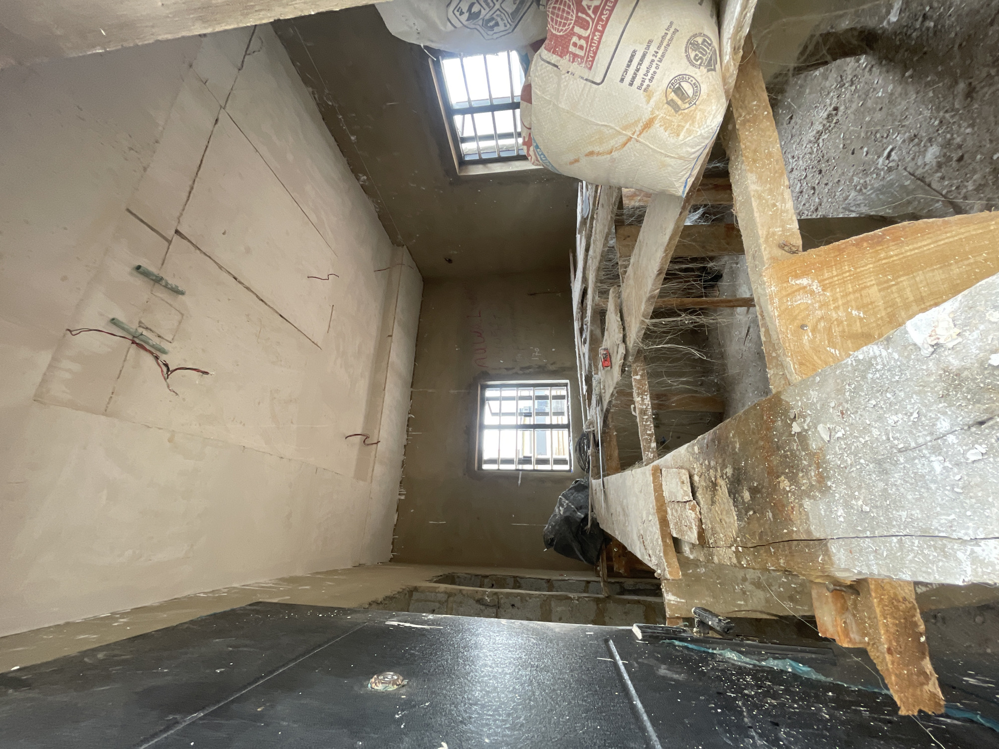 The image depicts an interior space under construction in what appears to be a residential or commercial building. Located in an undetermined area of Gosa, Nigeria, the room's walls are partially covered in a light-colored plaster or drywall compound. Electrical wiring is visible, protruding from the unfinished walls. Two windows with barred security grates are present, providing natural light. Scaffolding made of wooden planks dominates the right side of the frame, laden with two bags labeled "BUA GYPSUM PLASTER" and "PROUDLY NIGERIAN SUN," among other text. The floor is covered in black sheeting, possibly plastic or some other protective material. The overall impression is of a space undergoing active renovation or construction. No people are visible in the frame.
