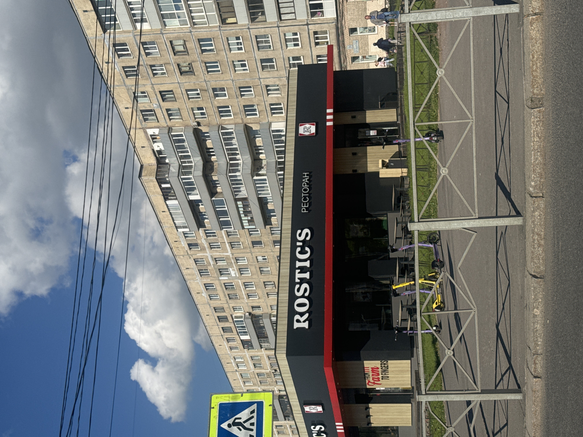 The image depicts an urban street scene in Russia during bright daylight, with a restaurant in the foreground and a large residential building behind it. The sky is blue with scattered white clouds, and power lines run across the upper left.
The main subject is a "ROSTIC'S" restaurant, identifiable by its prominent black and red sign with white lettering. Below "ROSTIC'S" is the Cyrillic word "РЕСТОРАН," meaning "RESTAURANT." The restaurant has an outdoor seating area with wooden tables and chairs, partially obscured by a metal barrier fence that separates it from the sidewalk and road. Several shared electric scooters, colored purple and yellow, are parked near the outdoor seating area. A small wooden sign near the restaurant entrance reads "FROM IT FARM TO FINGERS."
In the background, a large, multi-story residential building with numerous windows occupies the upper half of the image. Its facade appears to be light-colored, possibly concrete. Further back, another building has a sign with Cyrillic text, partially visible as "УЛИЦА" (Street) and "21".
On the far right, a paved road with a curb is visible. In the distance on the right, several people are walking on a sidewalk or path in front of buildings. The time of day appears to be sunny, creating distinct shadows from the fence and the restaurant building, suggesting either morning or late afternoon. A blue and yellow pedestrian crossing sign is partially visible in the bottom left corner.