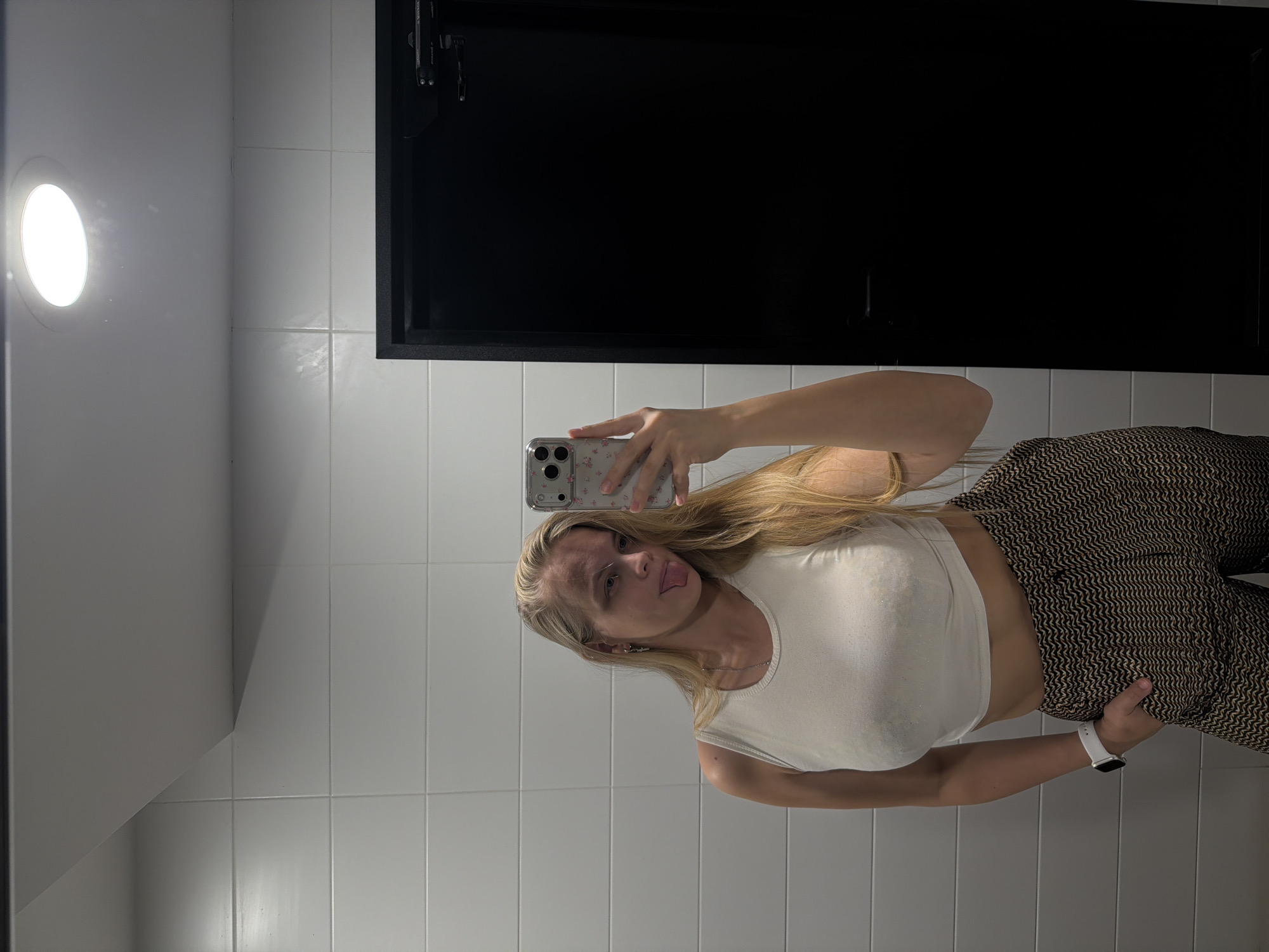 A woman is taking a selfie in what appears to be a bathroom. The bathroom has white tiles and a black fixture above her head. The woman is wearing a white crop top and patterned pants. She is holding a phone with a floral case, and she is wearing a white smartwatch. Her tongue is sticking out slightly. The presence of specific bathroom fixtures and the style of tiling are consistent with modern bathroom designs. The location cannot be determined to be in Izhevsk, Russia, solely based on the image.