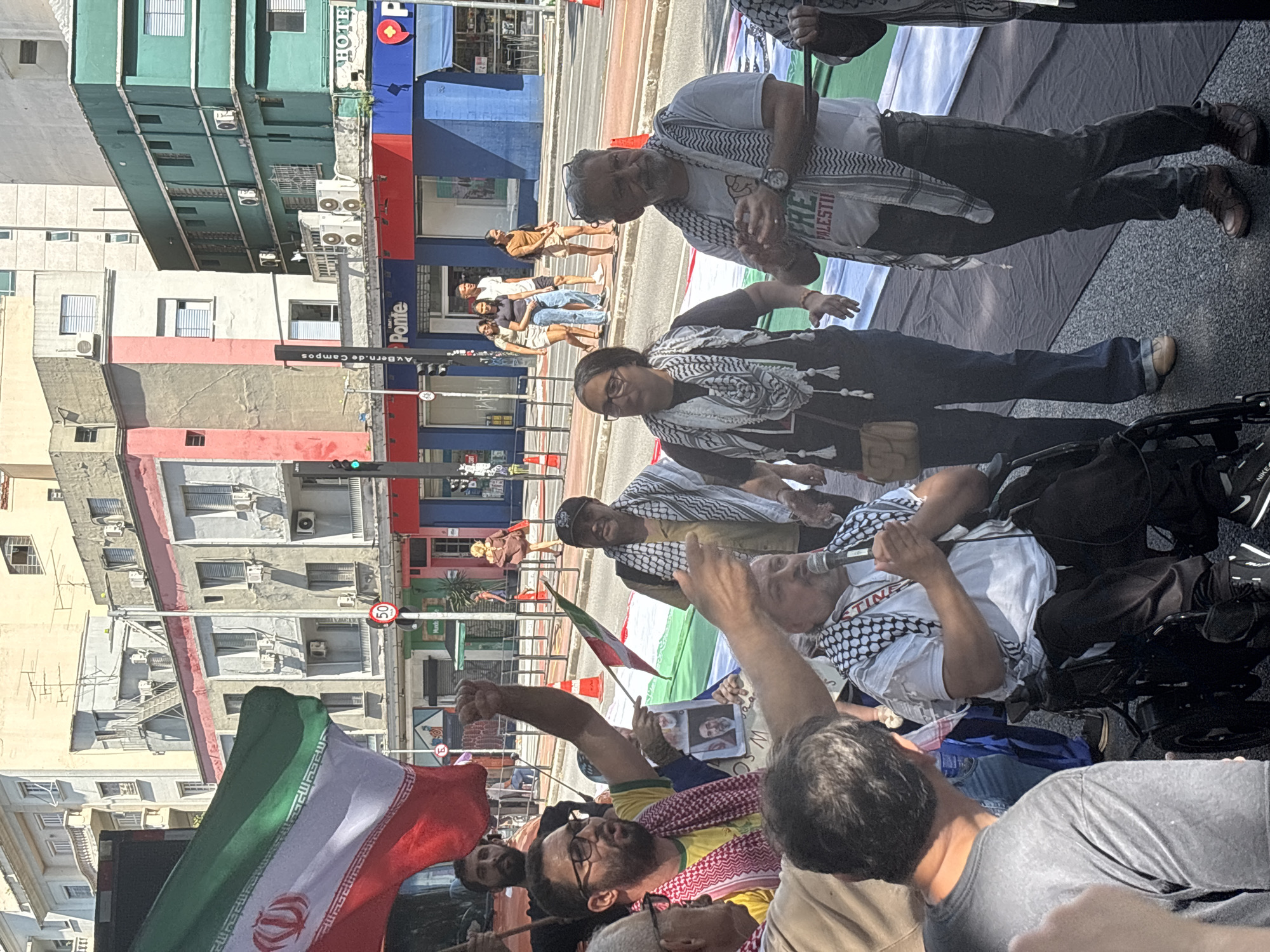The image depicts a protest in the city of São Paulo, Brazil. A group of people, some wearing keffiyehs, are gathered together. One person is speaking into a microphone, while others are holding an Iranian flag and a sign with portraits of people. Several buildings are visible in the background. A street sign indicating "Av. Bern. de Campos" is present, suggesting the location is Avenida Bernardino de Campos. The sky is sunny.