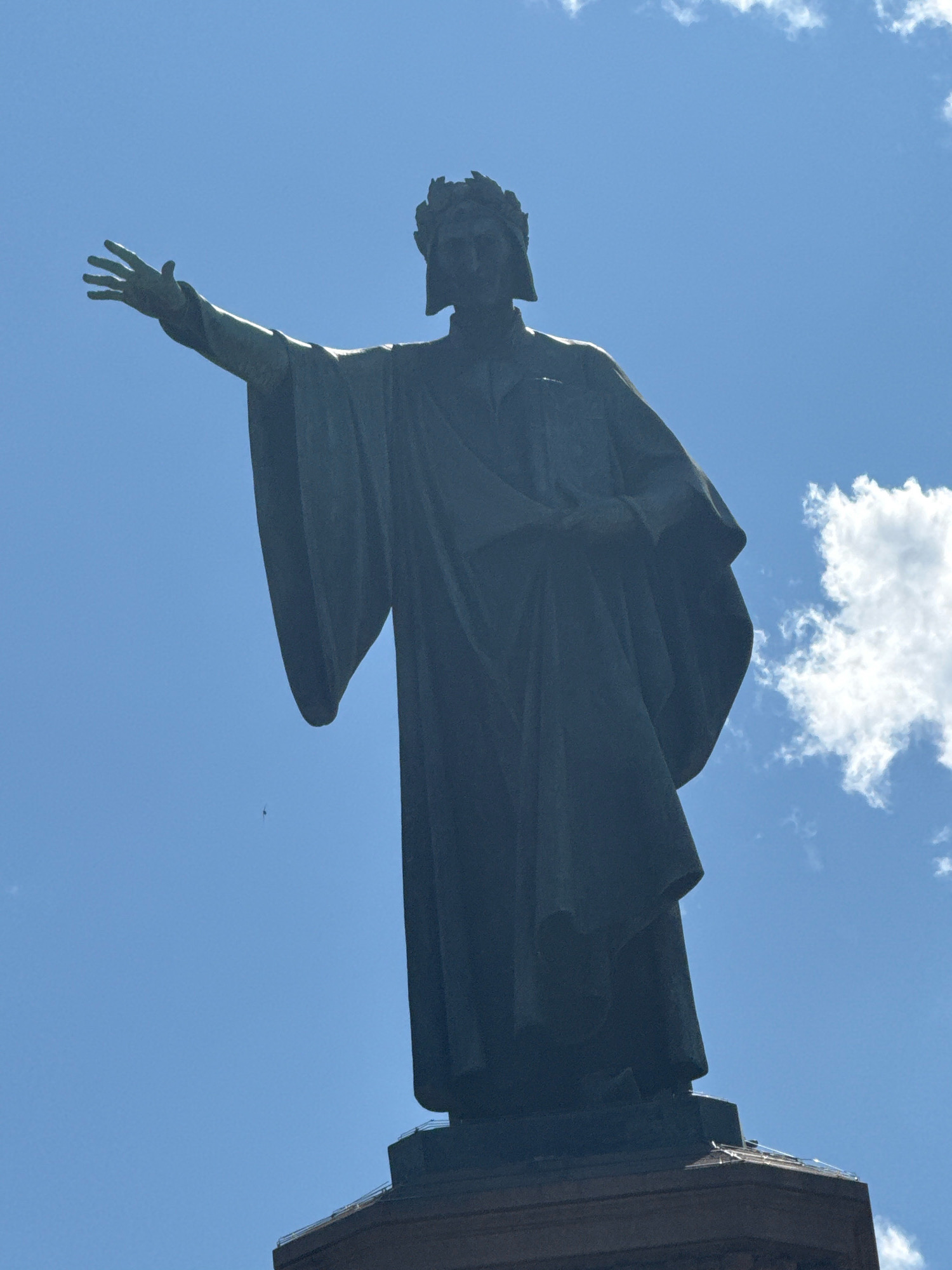 A dark bronze statue of Dante Alighieri, identifiable by his laurel wreath and traditional attire, stands against a bright blue sky with scattered white clouds. The statue is positioned on a pedestal. Dante's right arm is extended forward with his hand open, while his left arm is bent, holding a book close to his chest. A tiny black object, possibly a bird or an insect, is visible in the sky to the left of the statue's outstretched hand. The statue is located in Trento, Italy.