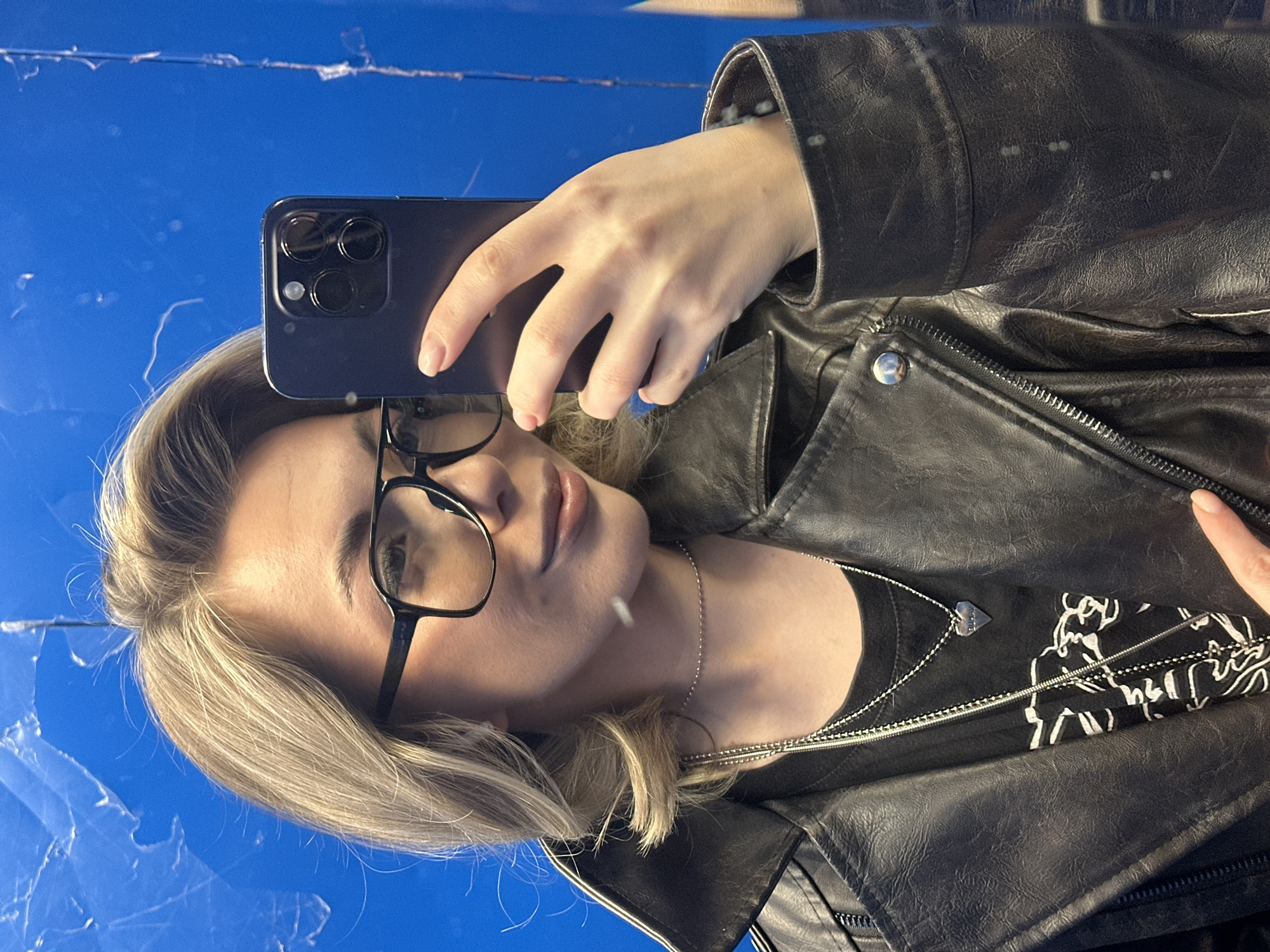 A woman with blonde hair and glasses is taking a selfie with a dark-colored smartphone. She is wearing a black leather jacket over a black shirt with a graphic design and a silver necklace with a heart pendant. The background is a bright blue color with some streaks or imperfections, suggesting it might be a wall or surface in a room. The photo has been rotated so the top of the frame is on the left. It is not possible to determine the specific location in Khimki, Russia, from the image alone.